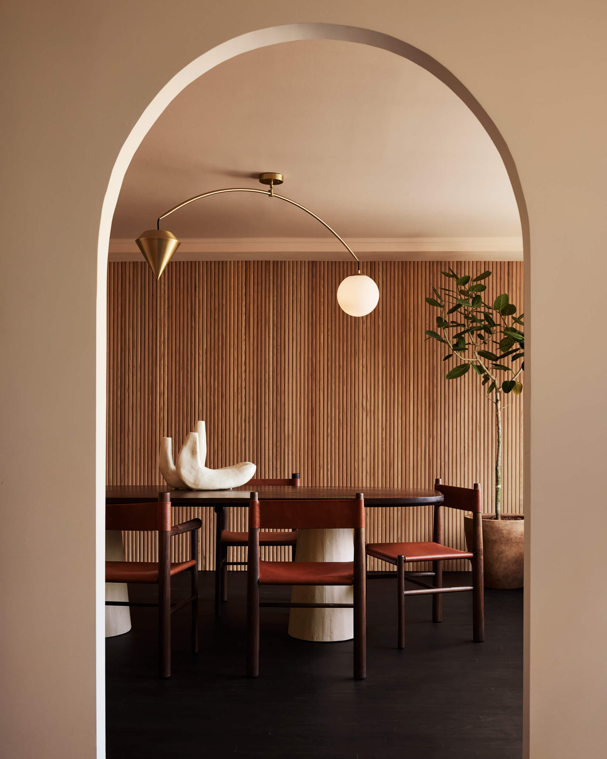 Arjé Home, North America — Minimalist dining room with arched entryway, wooden chairs, brass pendant lights, fluted wood paneling, and soft neutral palette.
