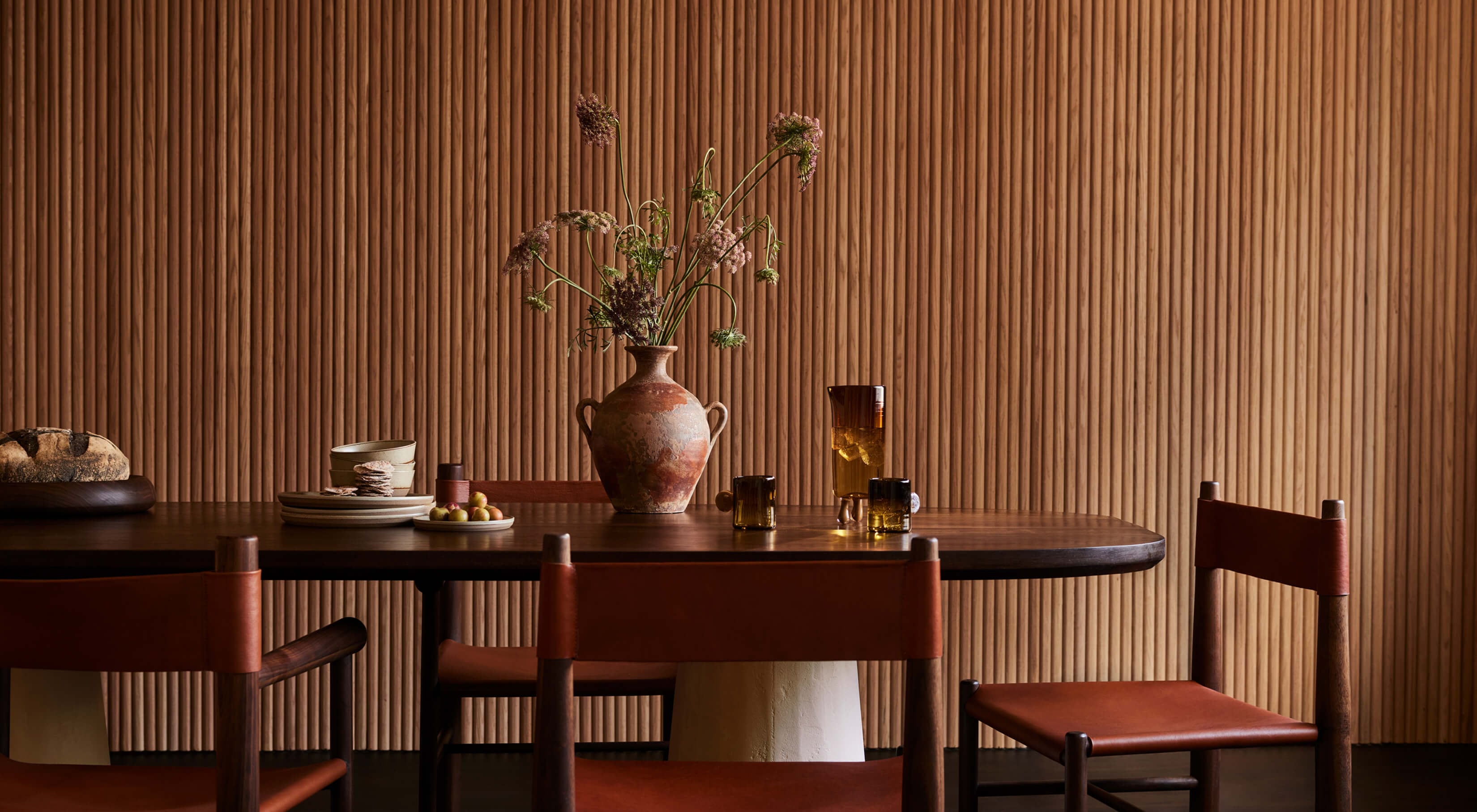 Arjé Home, North America — Walnut dining table with leather chairs, terracotta vessel with dried botanicals, corrugated ochre wall paneling, warm ambient light.