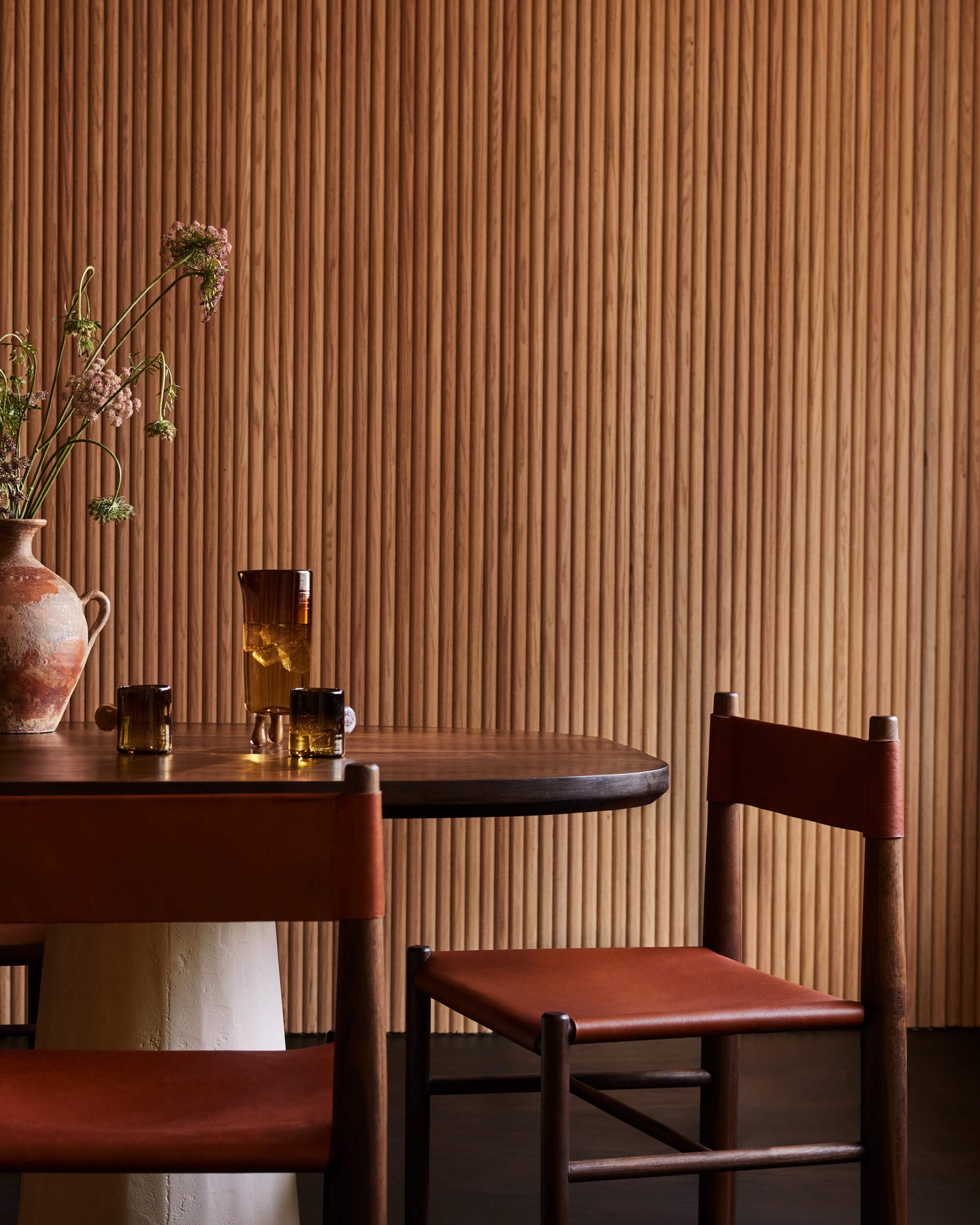 Arjé Home, North America — Minimalist dining room with cognac leather chairs, dark wood round table, amber glassware, dried florals in terracotta vase, and warm corrugated wall paneling.