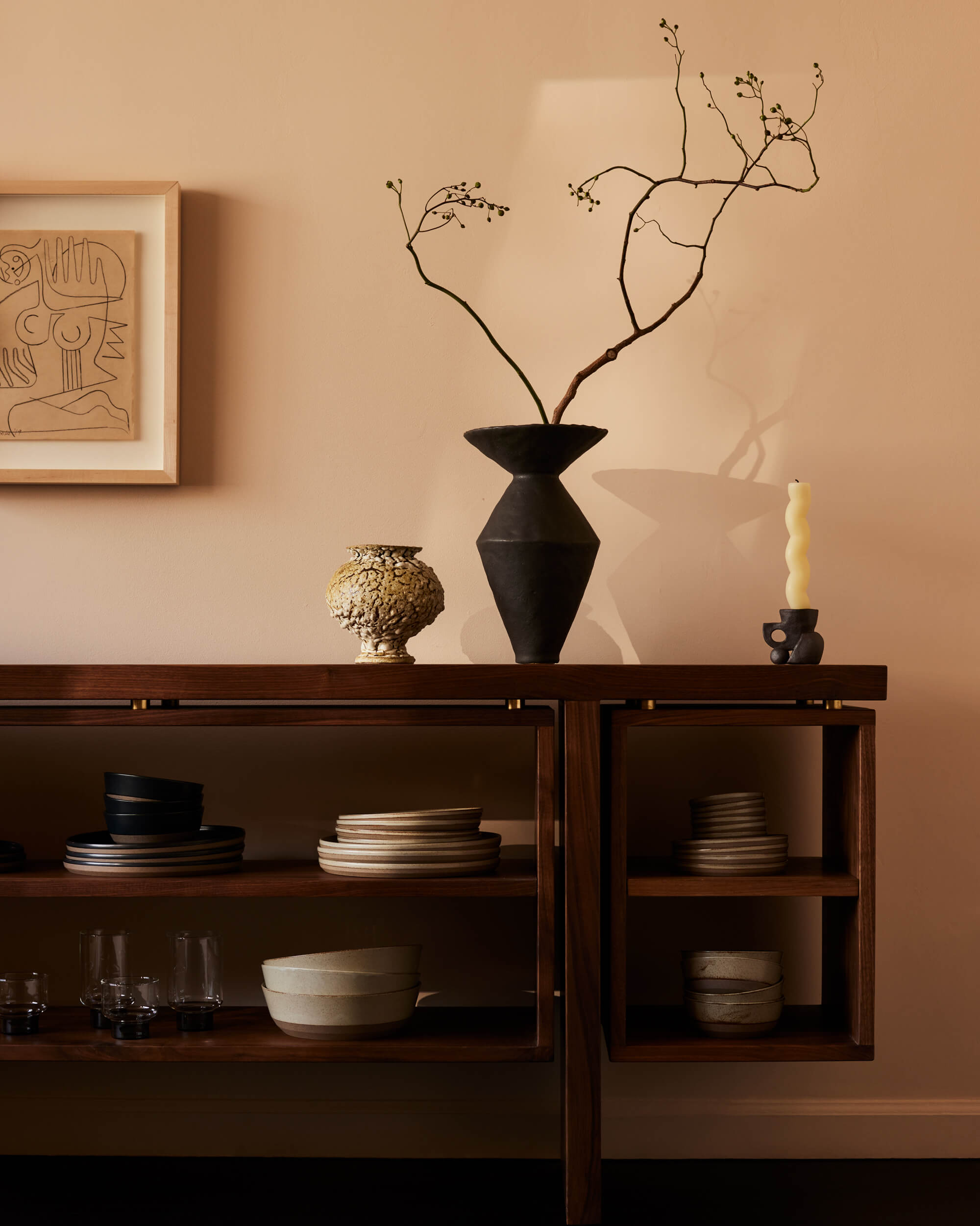 Arjé Home, North America — Minimalist dining sideboard with sculptural black vase, dried botanicals, gold textured vessel, and stacked dinnerware against warm neutral wall.