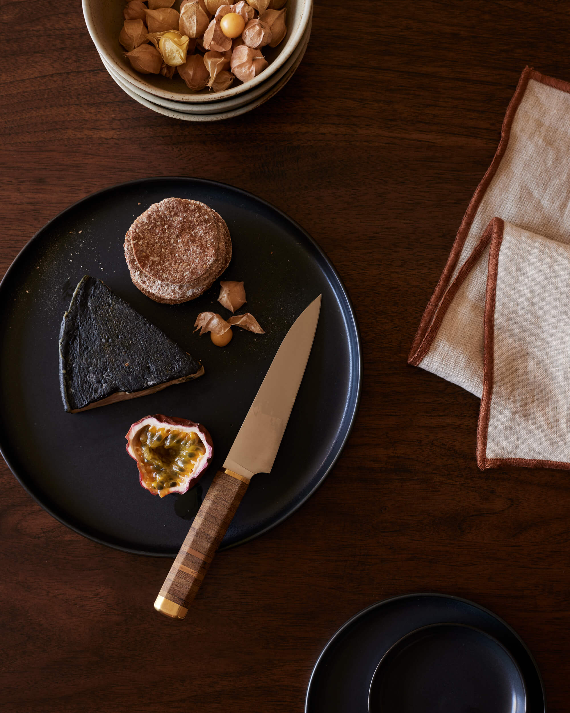 Arjé Home, North America — Dark ceramic plate with chocolate desserts, passion fruit, sugar-dusted cake, and wooden knife on rich wood table with linen napkin.