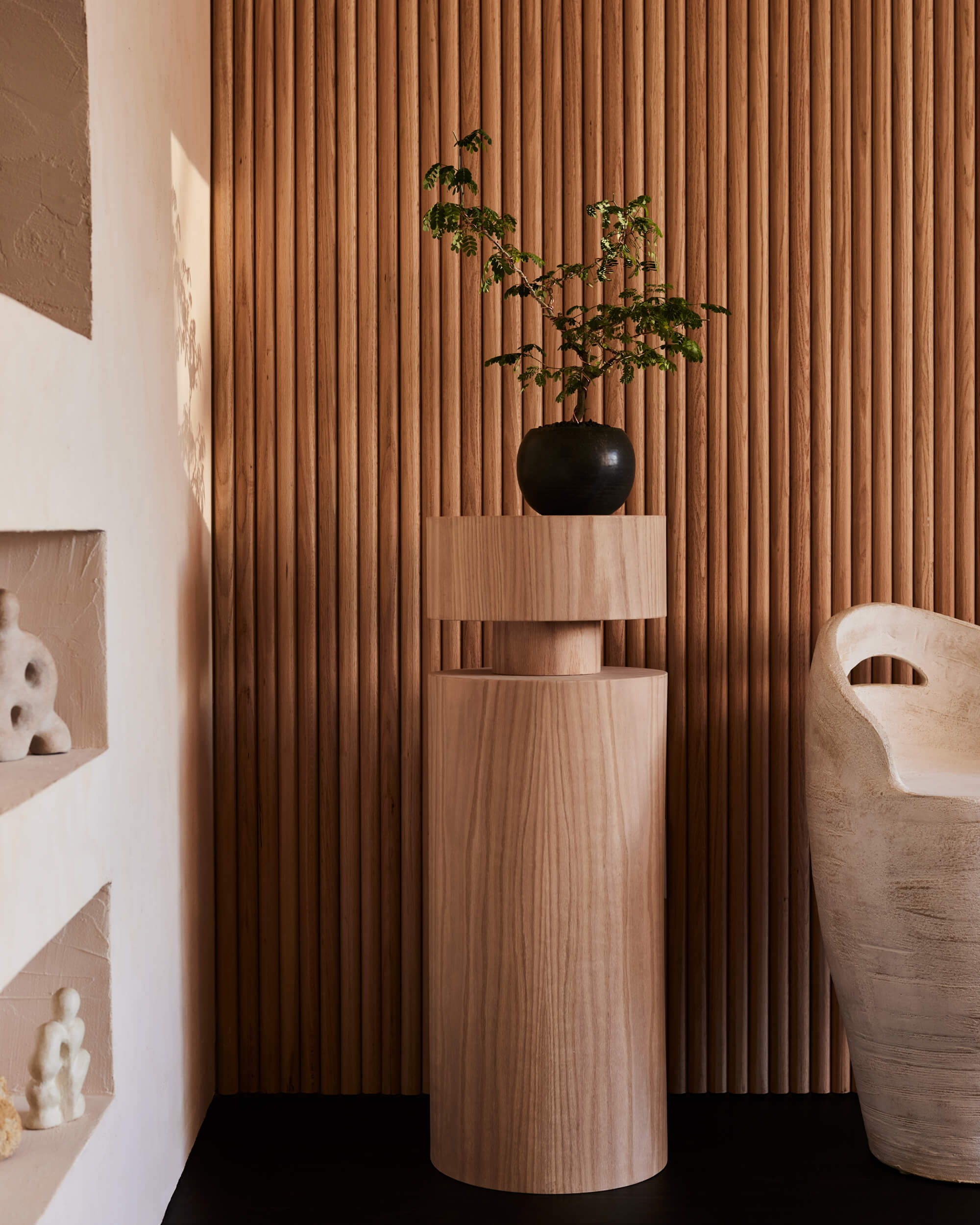 Arjé Home, North America — Wooden pedestal with black planter and greenery against fluted wood wall, minimalist interior design.