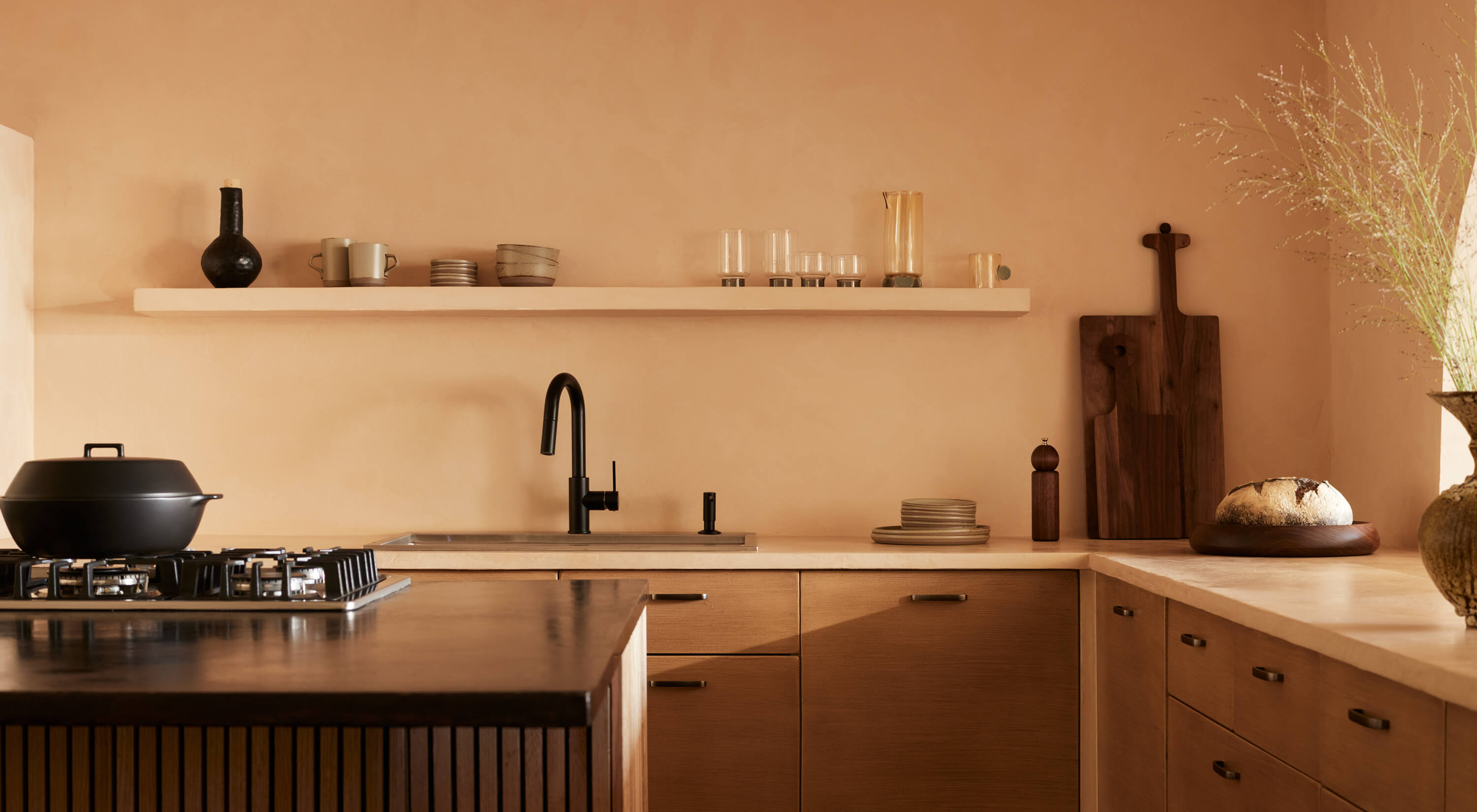 Arjé Home, North America — Minimalist kitchen with terracotta walls, natural wood cabinetry, black matte fixtures, and curated ceramic vessels on floating shelf.