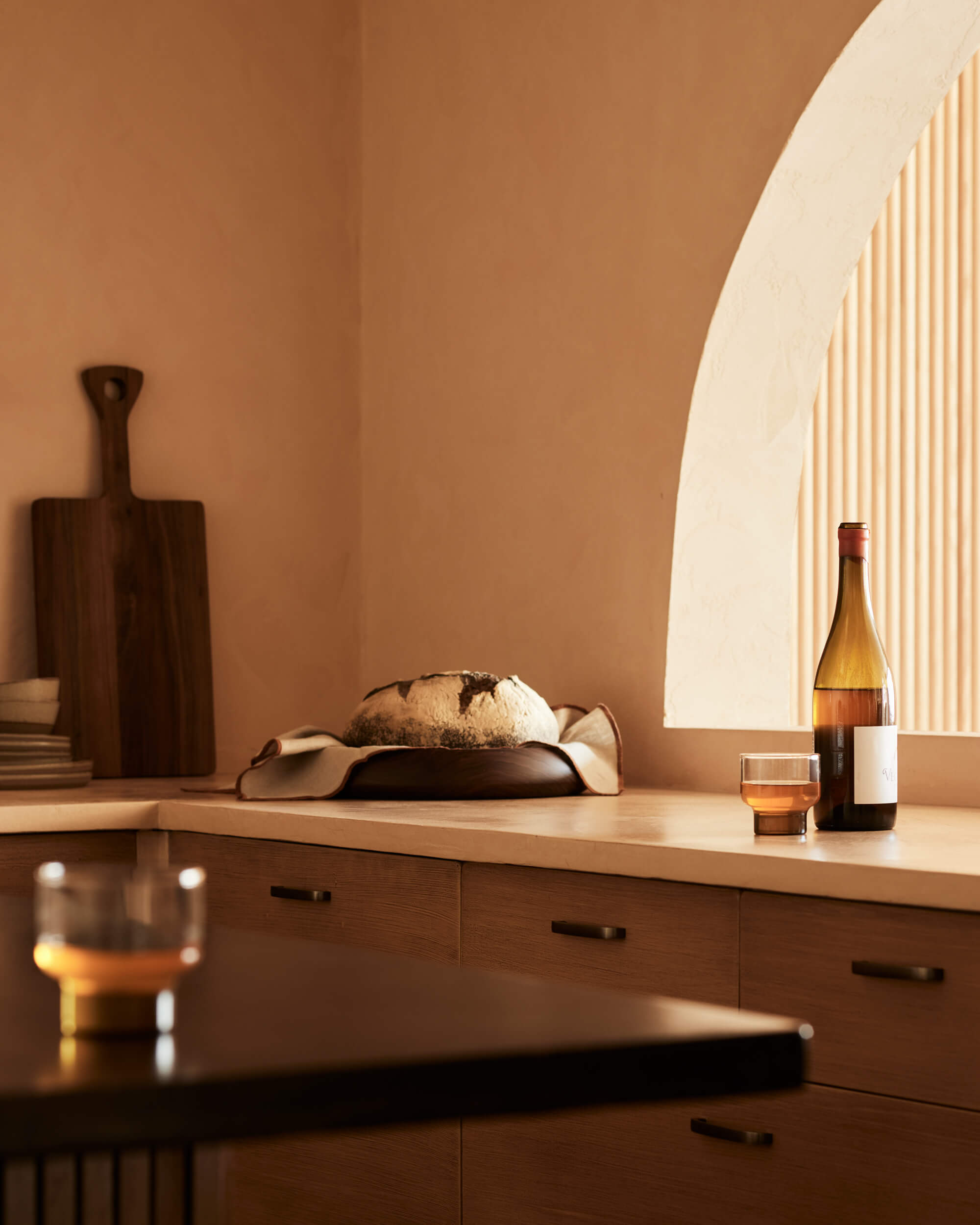 Arjé Home, North America — Minimalist kitchen counter with bread, wine bottle, wooden cutting board, and warm natural light through curved archway.