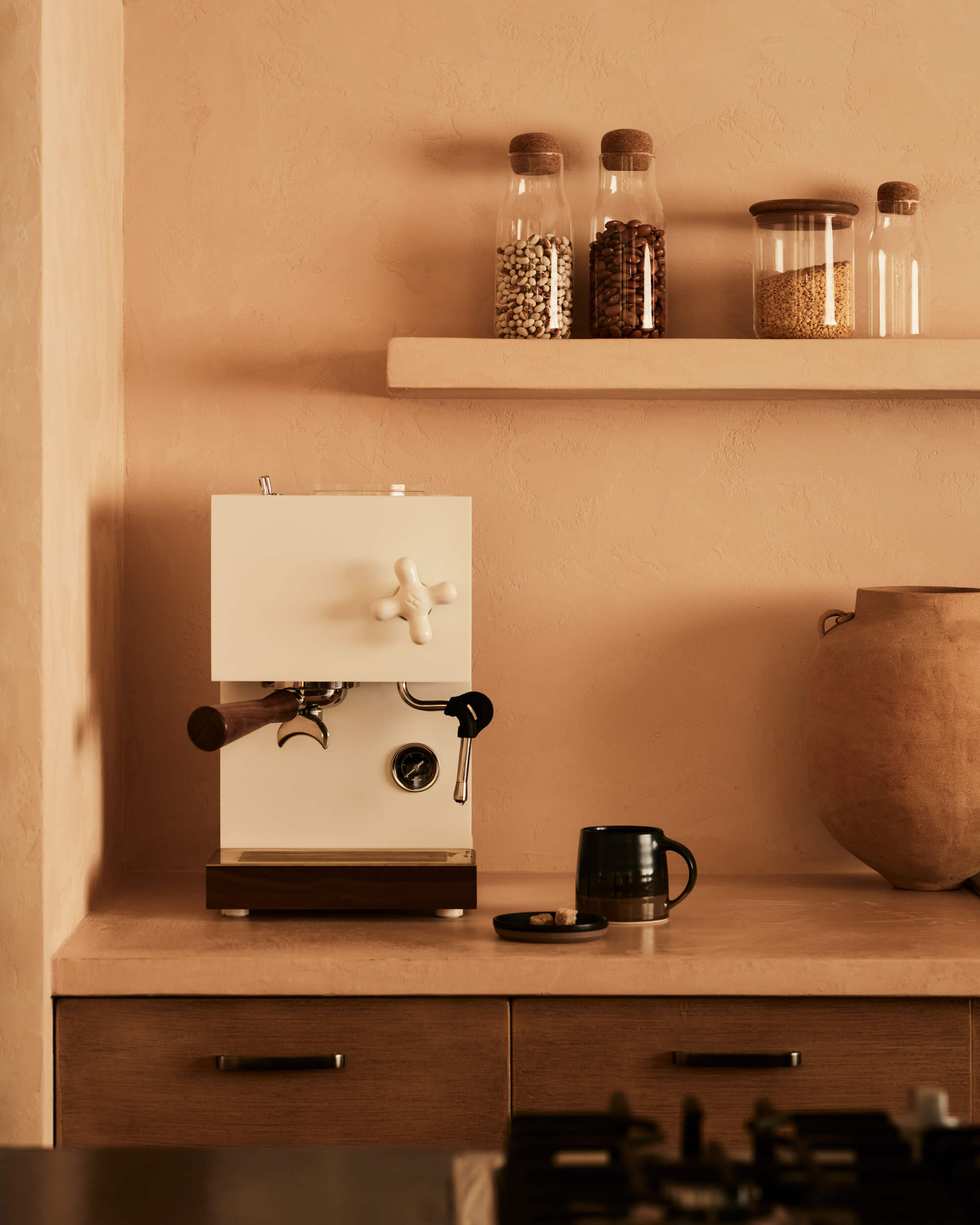 Arjé Home, North America — Minimalist kitchen counter with cream espresso machine, glass storage jars, terracotta vessel, and warm monochromatic aesthetic.