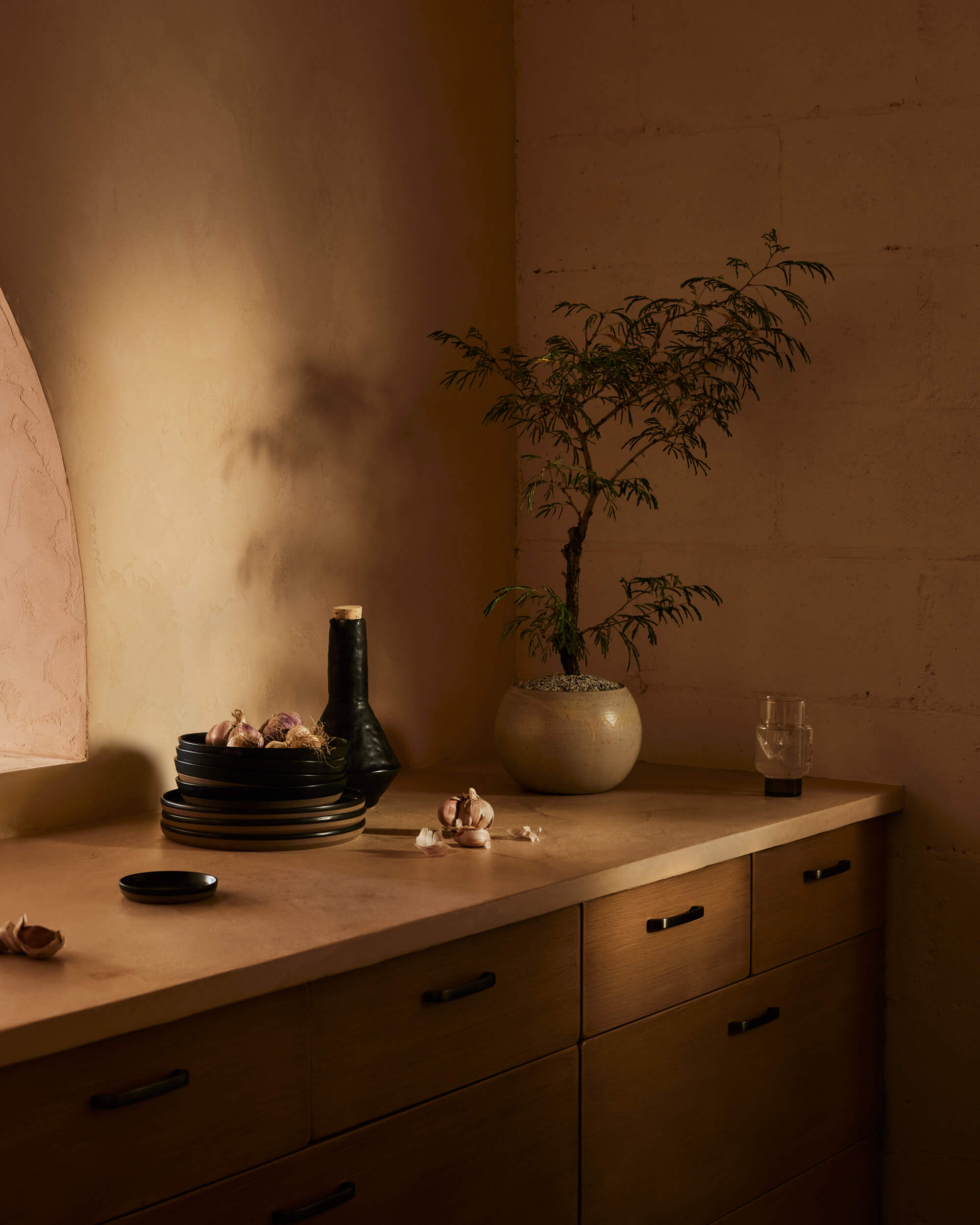 Arjé Home, North America — Minimalist wooden dresser with stacked black dinnerware, potted plant, and garlic cloves against warm monochromatic wall with soft directional light.