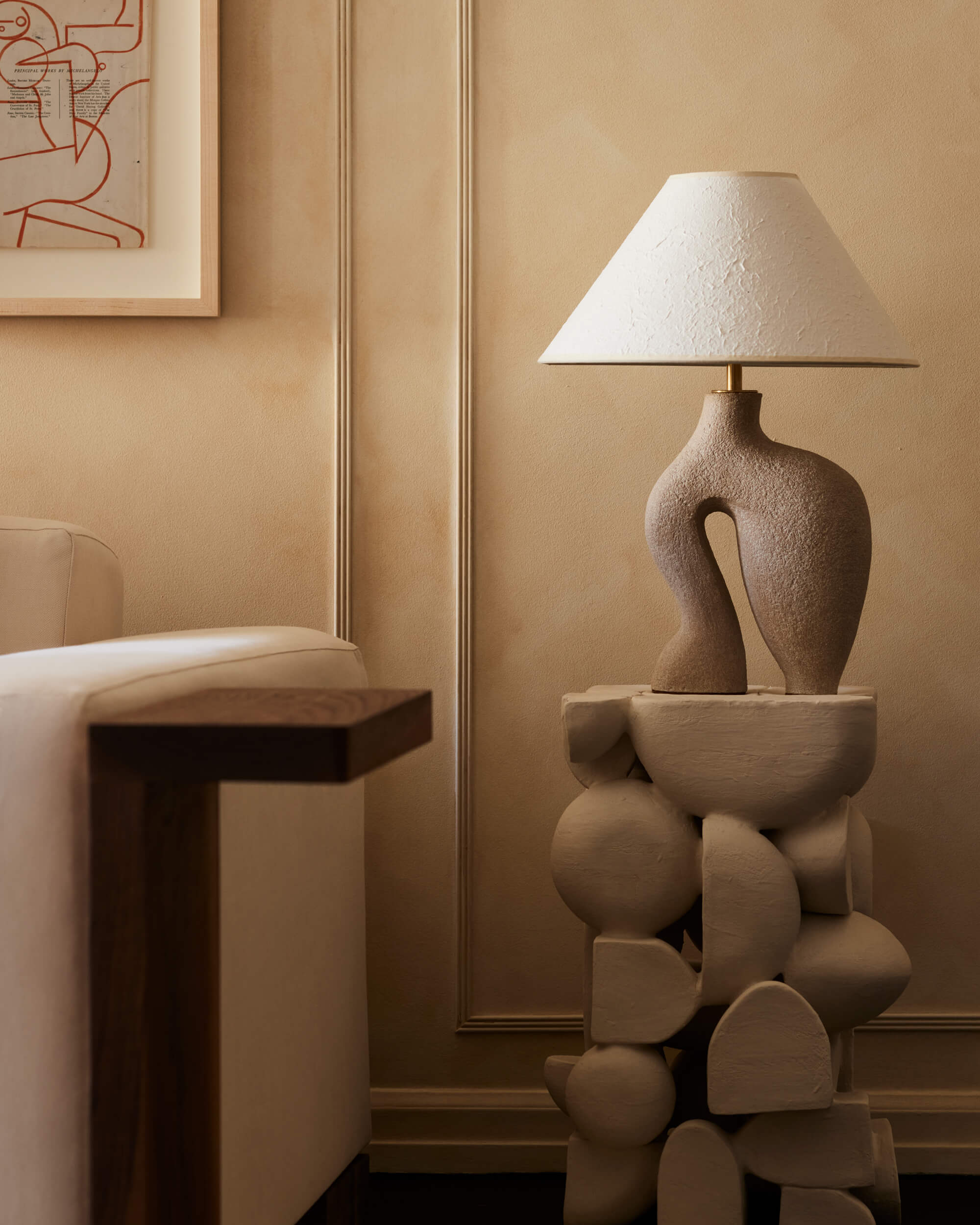 Arjé Home, North America — Sculptural stacked ceramic base table lamp with linen shade in monochromatic earth-tone interior with paneled wall.