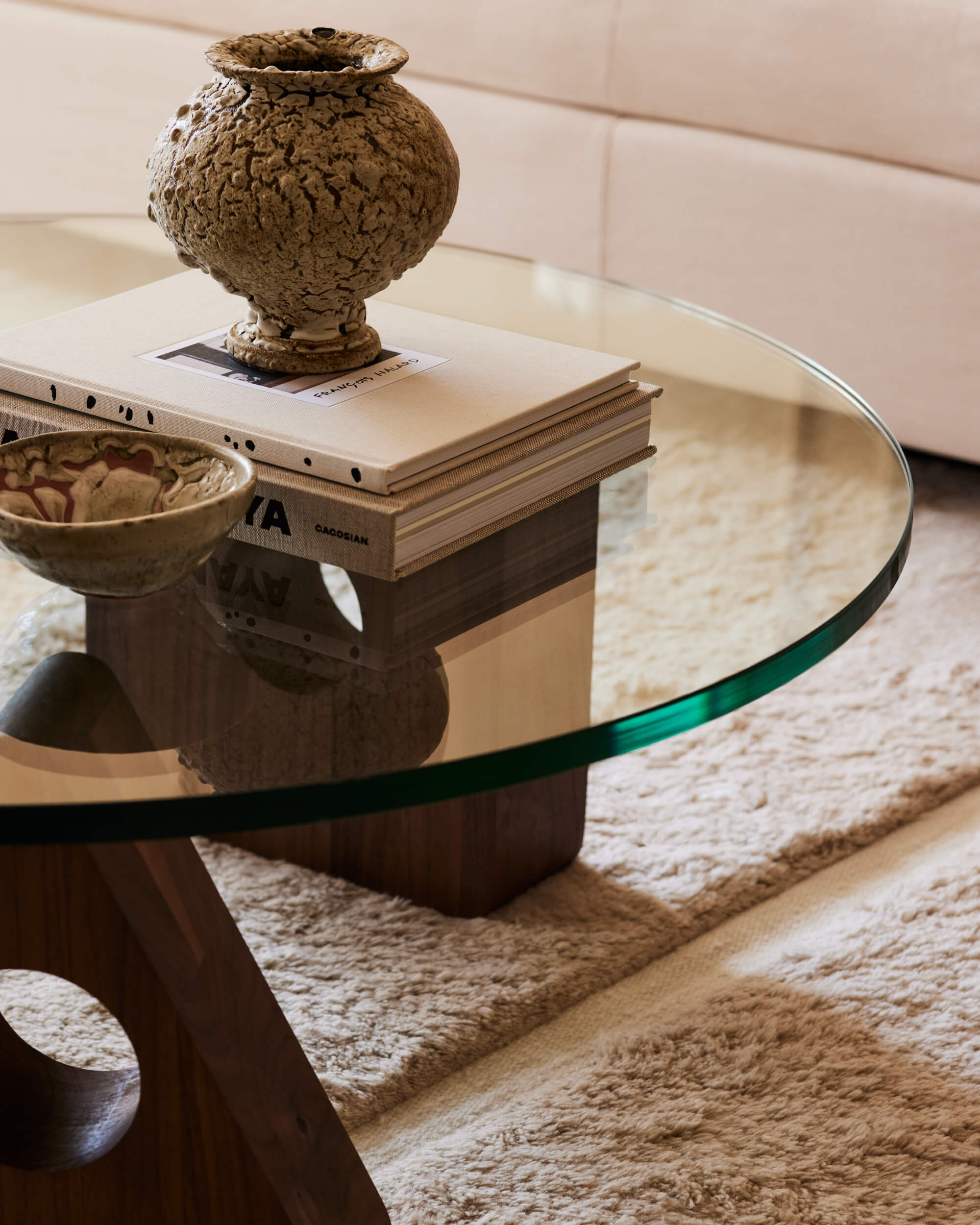 Arjé Home, North America — Sculptural terracotta vessel on stacked books atop round glass table with carved wood base, neutral textiles and soft natural light.