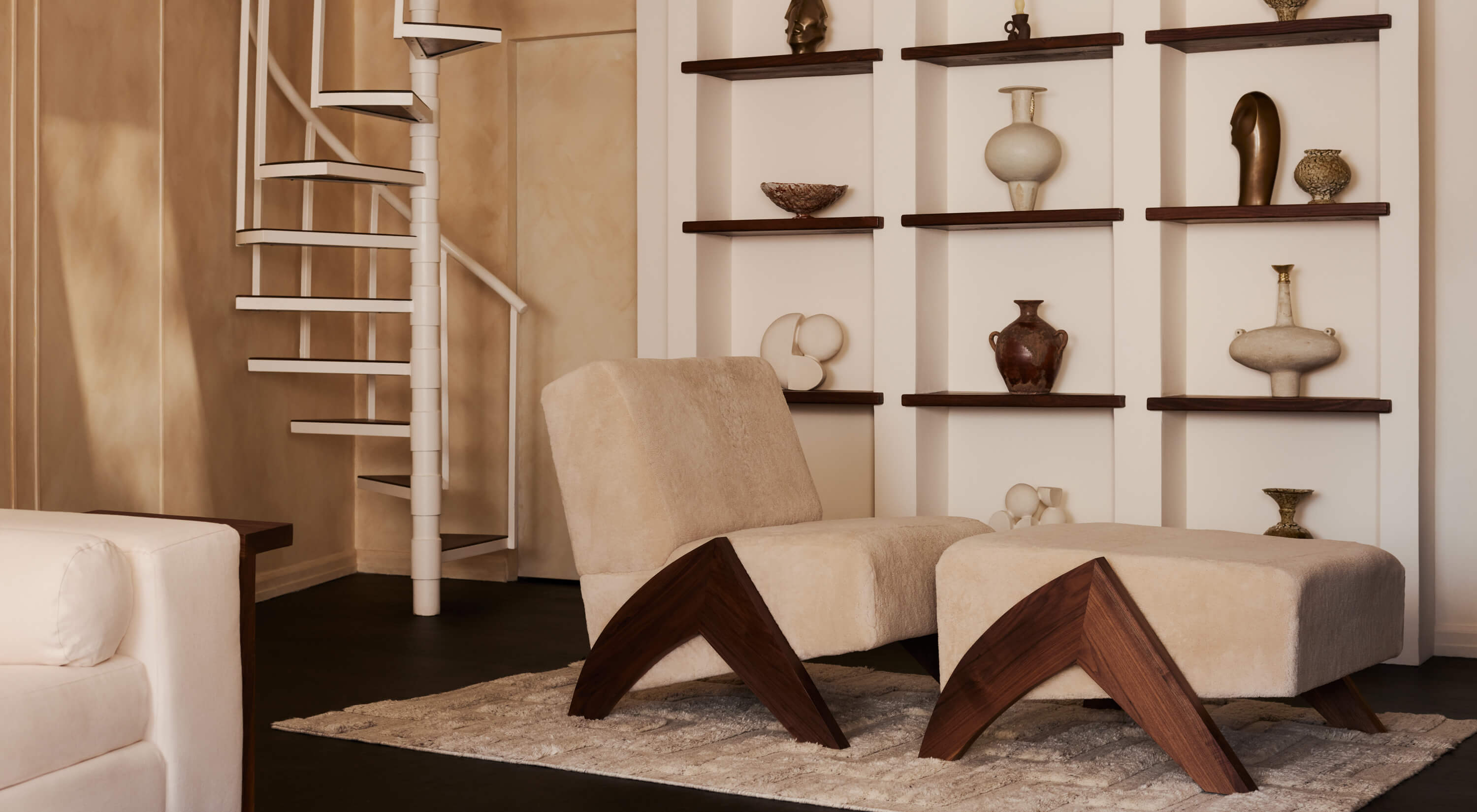 Arjé Home, North America — Cream upholstered lounge chairs with walnut frames on stone terrazzo floor, dark wood shelving displaying sculptural vessels and ceramics, spiral staircase in background.