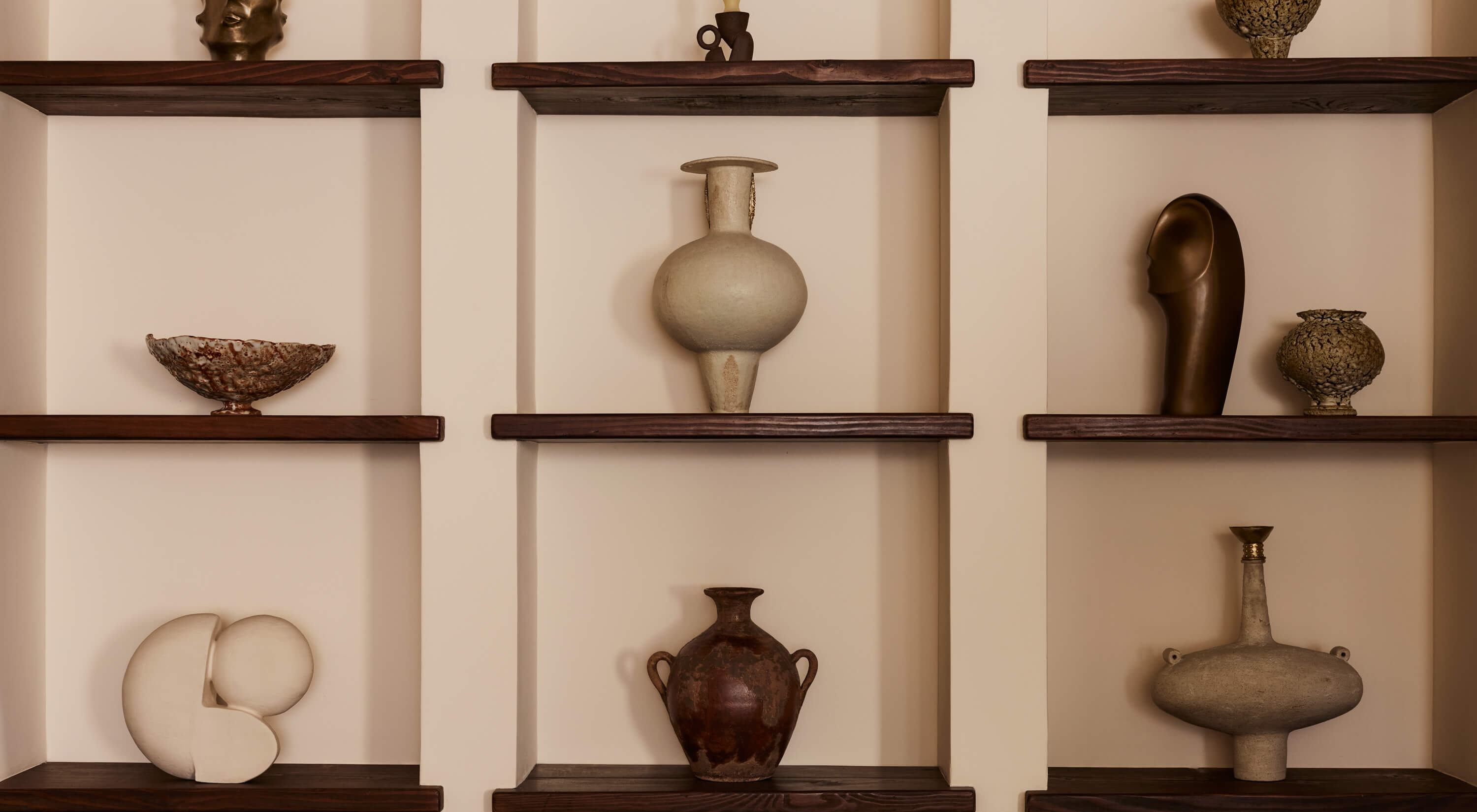 Arjé Home, North America — Dark wooden shelves display curated ceramic vessels and sculptural objects against cream walls, architectural display design for luxury hospitality interiors.