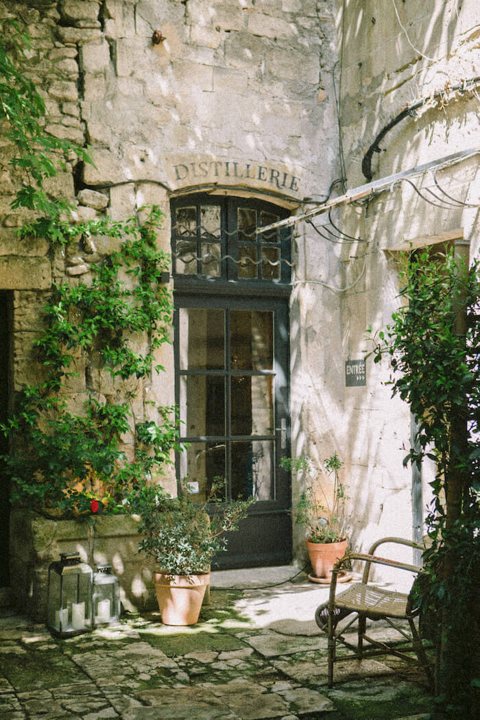 Weathered stone courtyard entrance to a distillery in Arles, with ivy-clad walls and terra-cotta planters casting afternoon shadows.