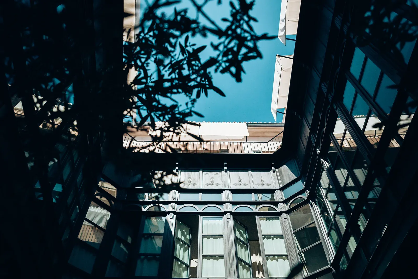 Sant Francesc Hotel Singular, Balearic Islands, Spain — Courtyard viewed from above with classical columns, tall windows, and dappled foliage shadows against bright blue sky.