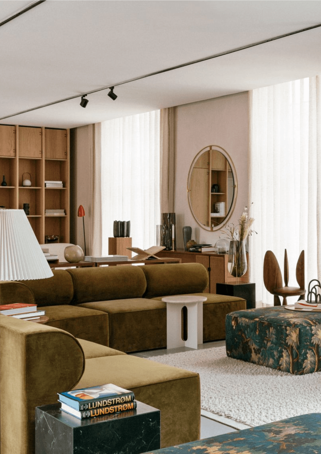 The Audo, Europe — Warm-toned living room with olive velvet sectional, wooden shelving, oval mirror, and natural light from floor-to-ceiling windows.