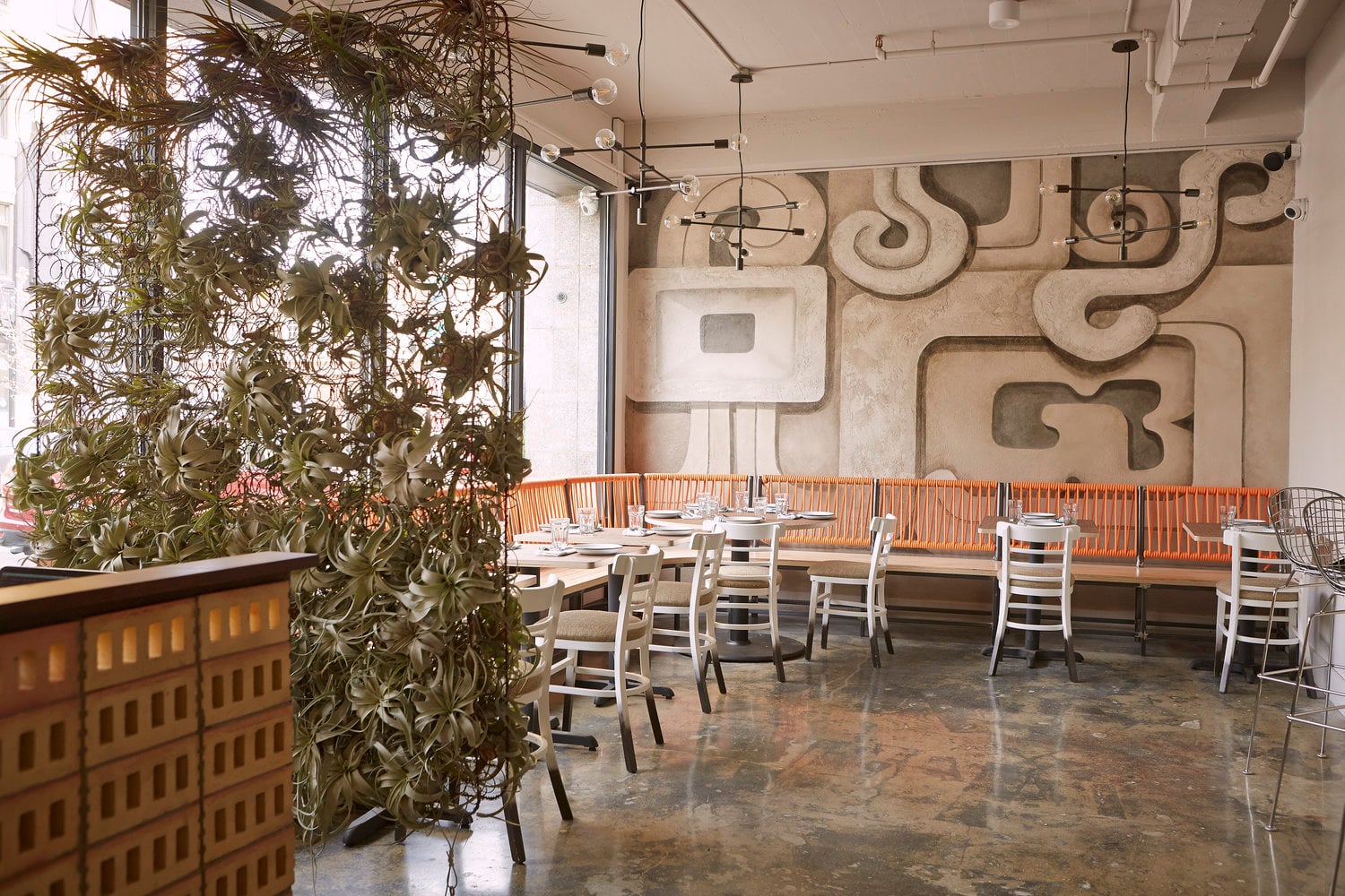 Barrio Costero, North America — Modern restaurant dining room with concrete floors, climbing ivy partition, sculptural wall art, pendant lighting, and orange-upholstered banquette seating.