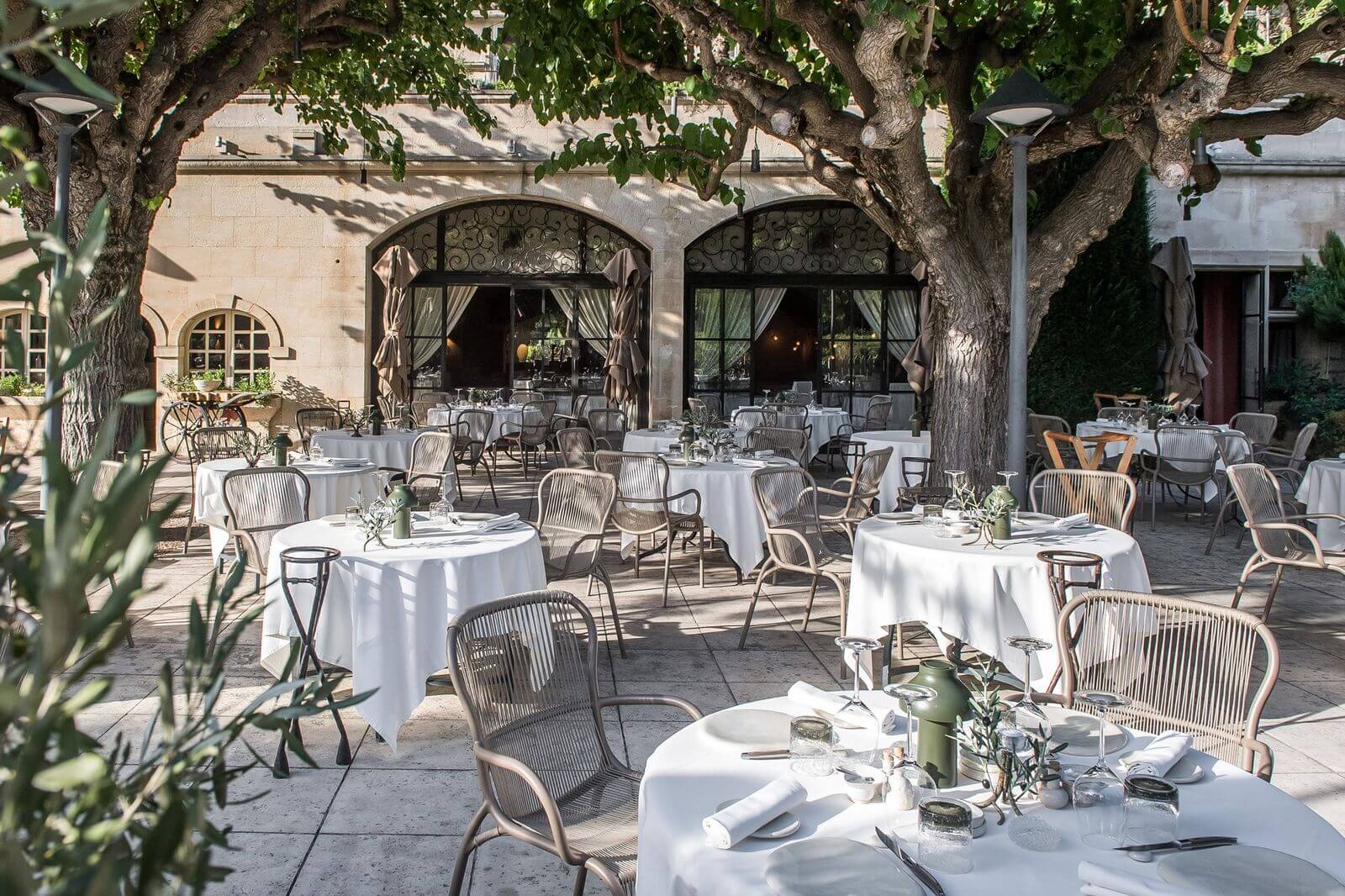 Baumanière, Europe — Ochre stone courtyard dining terrace shaded by mature plane trees, white-clothed tables, woven chairs, ornate arched windows, Mediterranean noon light.