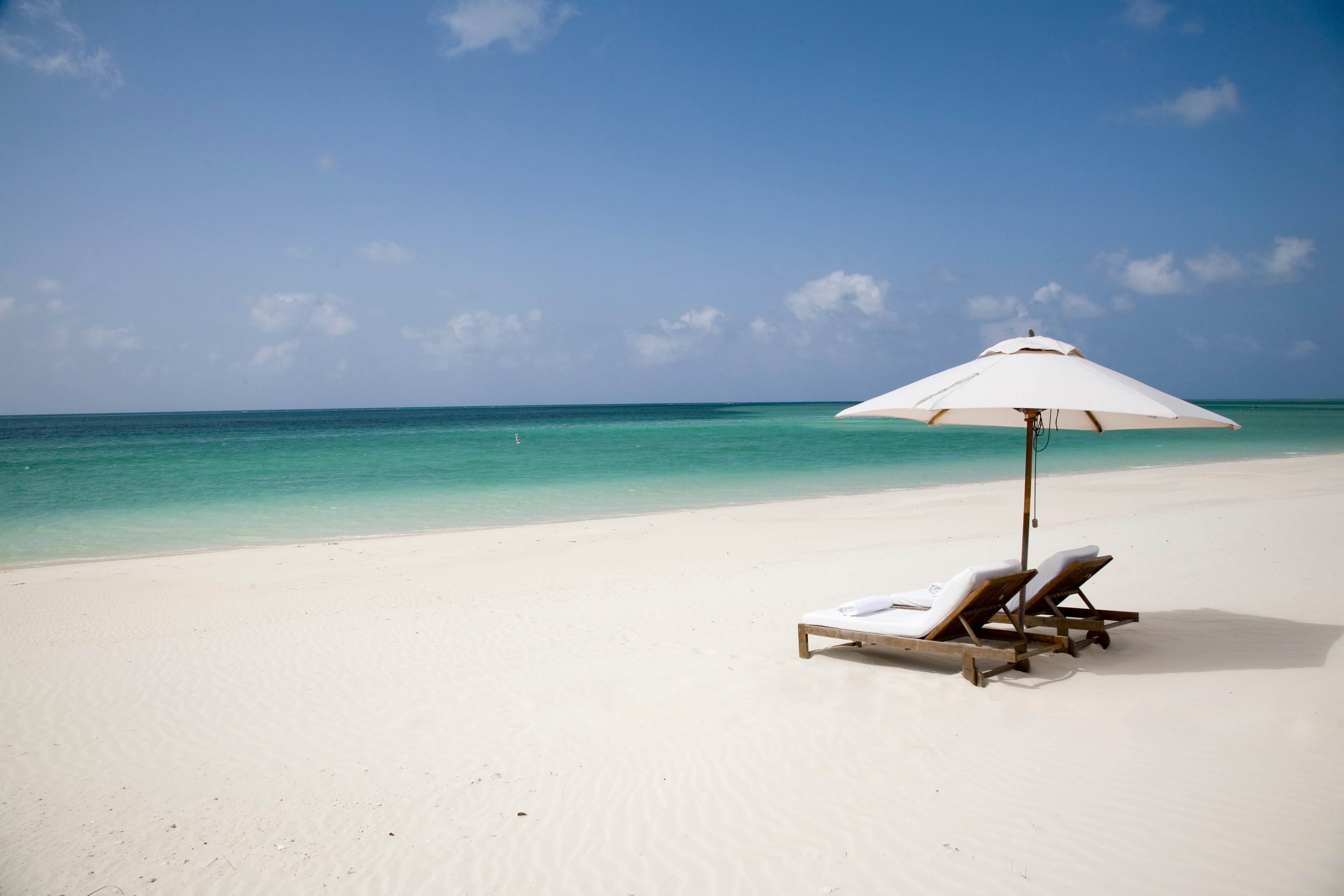 COMO Parrot Cay, North America — Two wooden loungers with white cushions and umbrella on pristine sand facing turquoise ocean under clear sky.