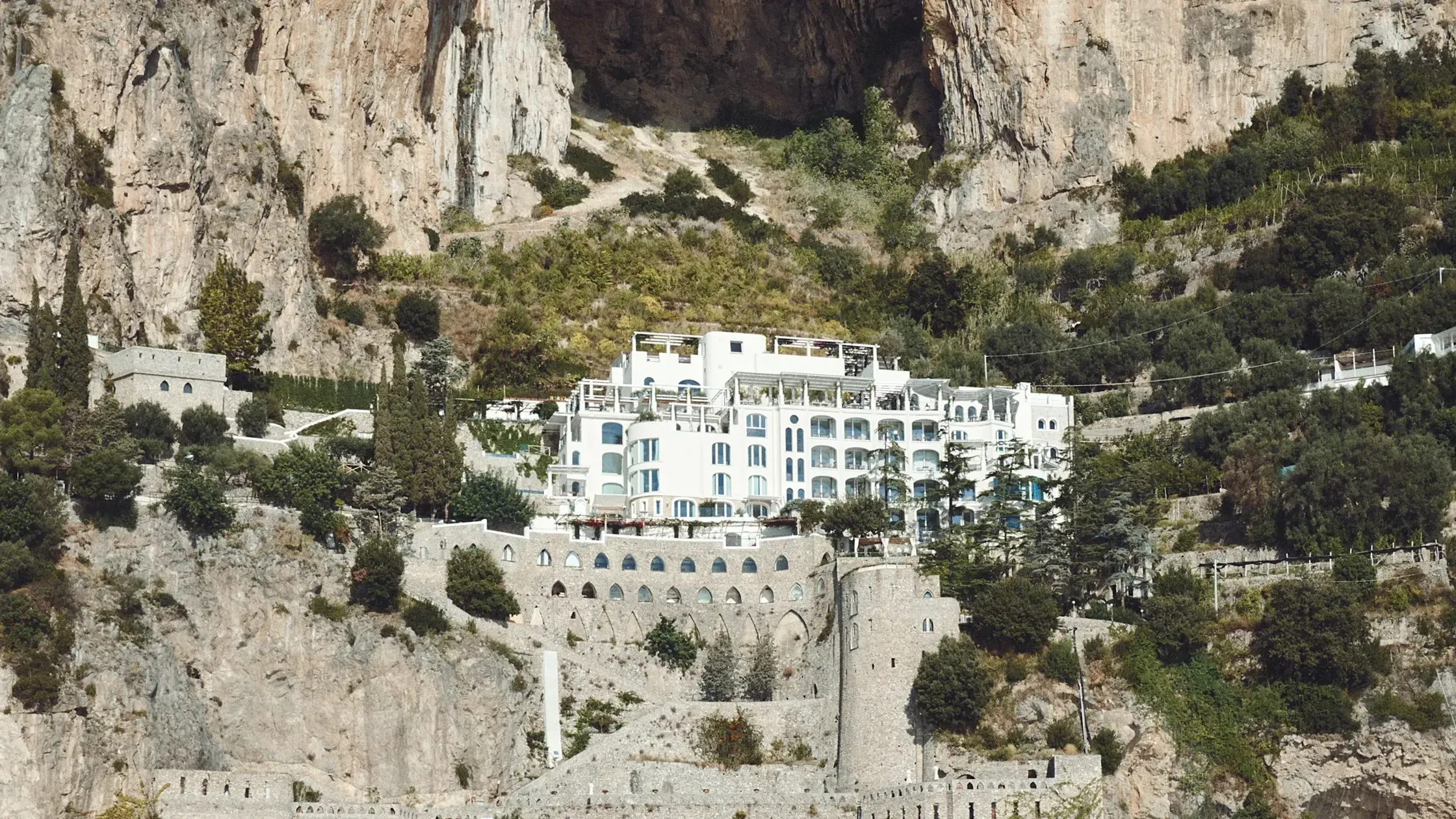 Borgo Santandrea, Italy — whitewashed luxury hotel nestled into limestone cliffs with cascading terraces, stone fortifications, and Mediterranean vegetation in dramatic seaside setting.