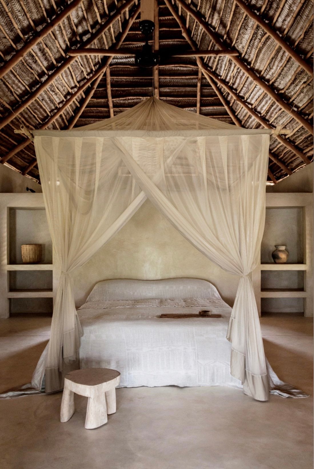 Sussurro, Africa — Minimalist canopy bed with sheer linen curtains in whitewashed bedroom beneath woven thatch ceiling, flanked by built-in alcoves.