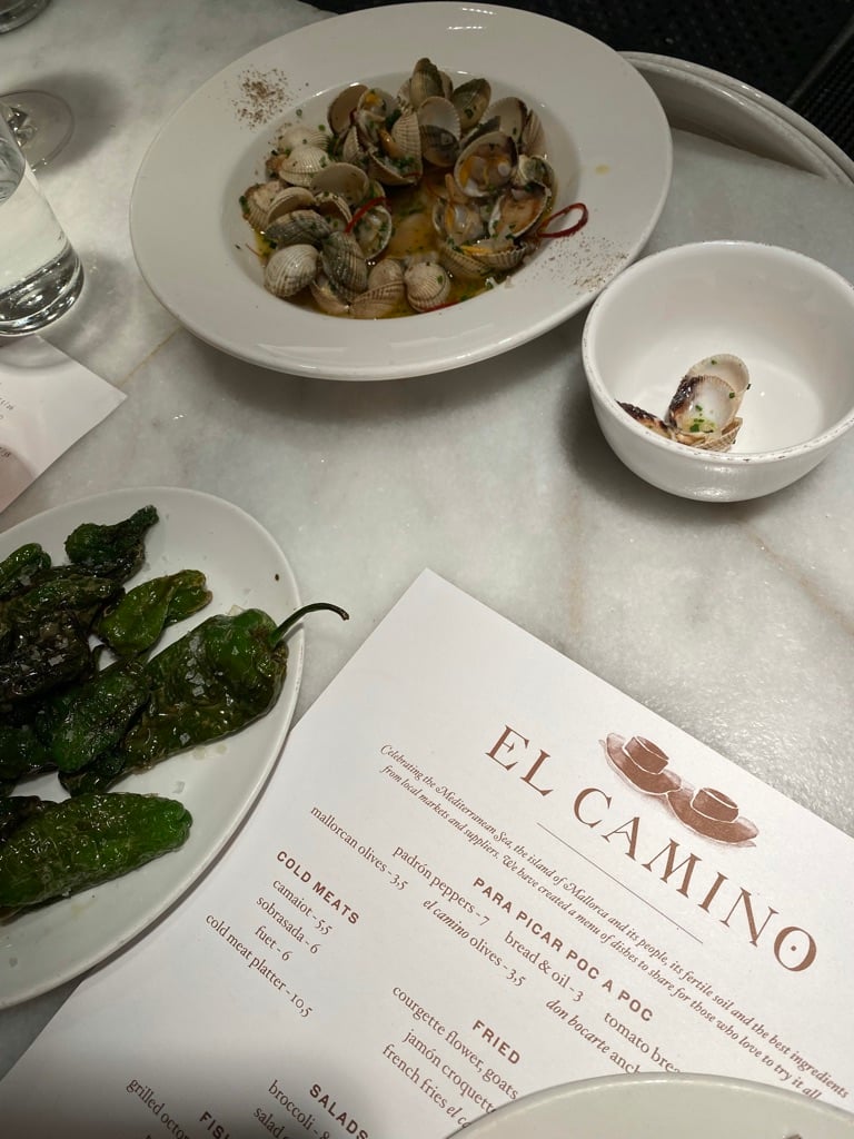 El Camino, Balearic Islands, Spain — clams in white wine sauce, charred peppers, and menu on marble table at coastal seafood restaurant.