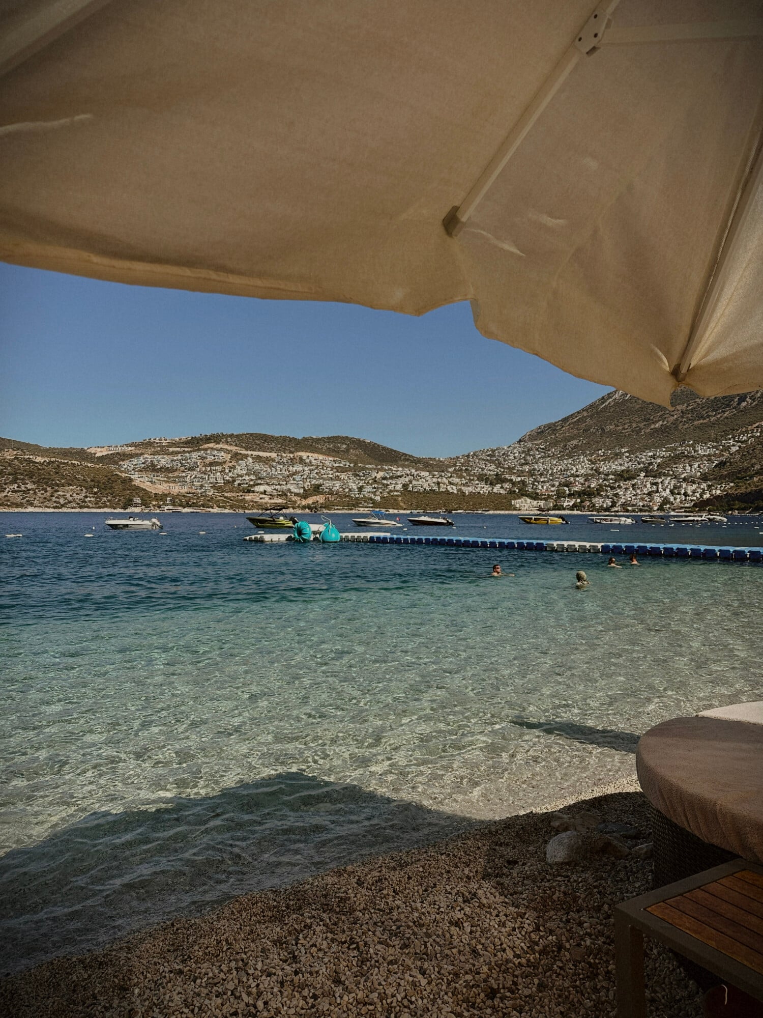 Kalkan Beach Park Club, Asia — Beachfront cove with turquoise water, pebbly shore, umbrellas, anchored boats, and terraced hillside architecture under clear sky.