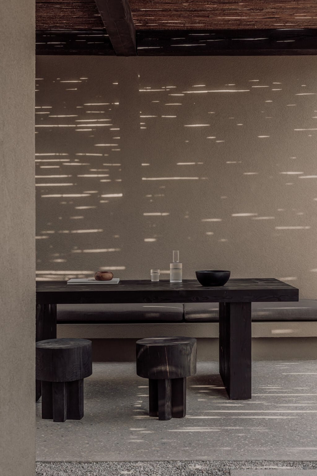 Casa Cook Samos, Greece — Minimalist dining space with dark wood table and stools, concrete floor, sunlight casting linear shadows on textured wall.