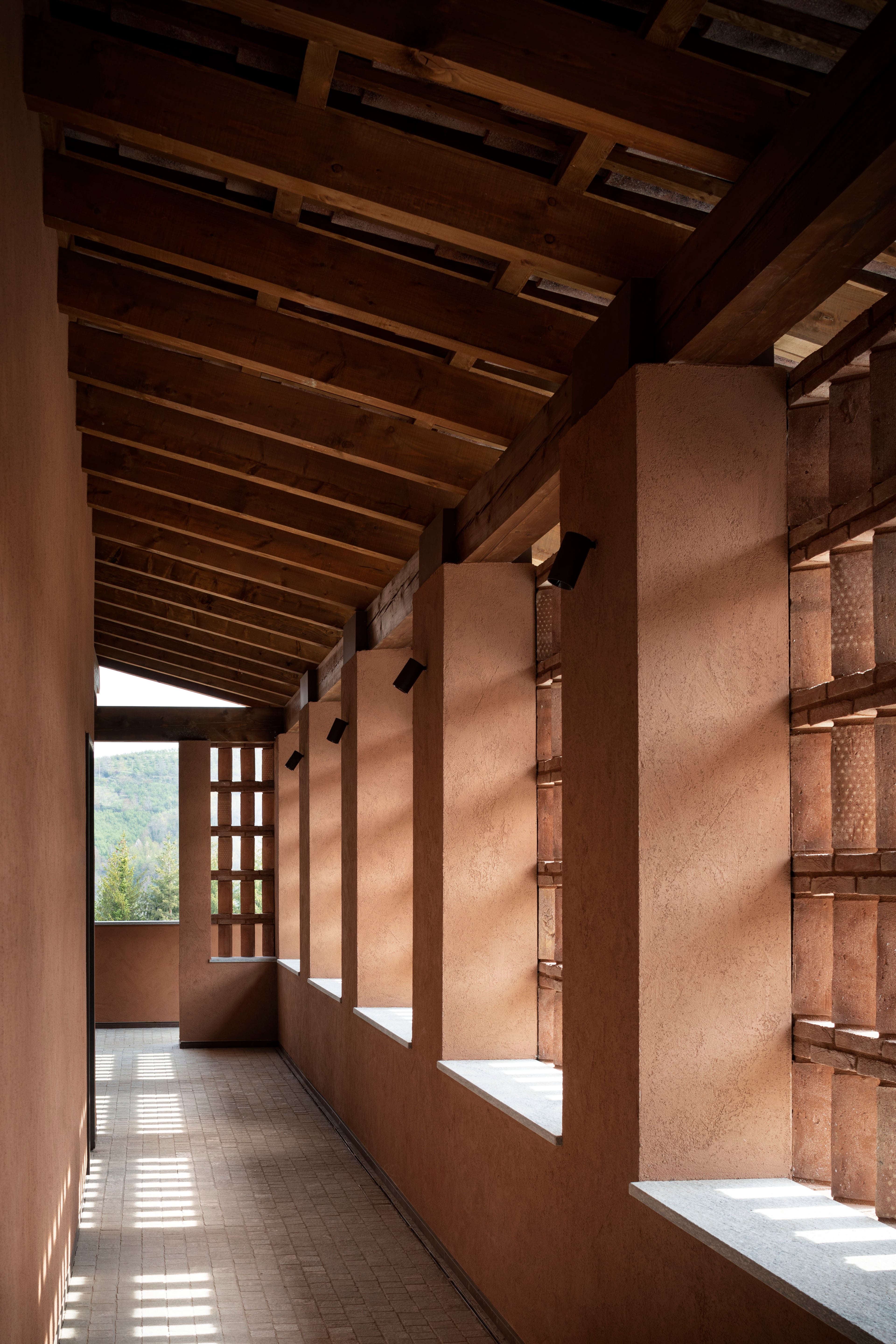 Casa di Langa, Europe — Terracotta corridor with exposed wooden beam ceiling, stone pillars, and slatted window screens casting geometric light patterns across tiled floor.