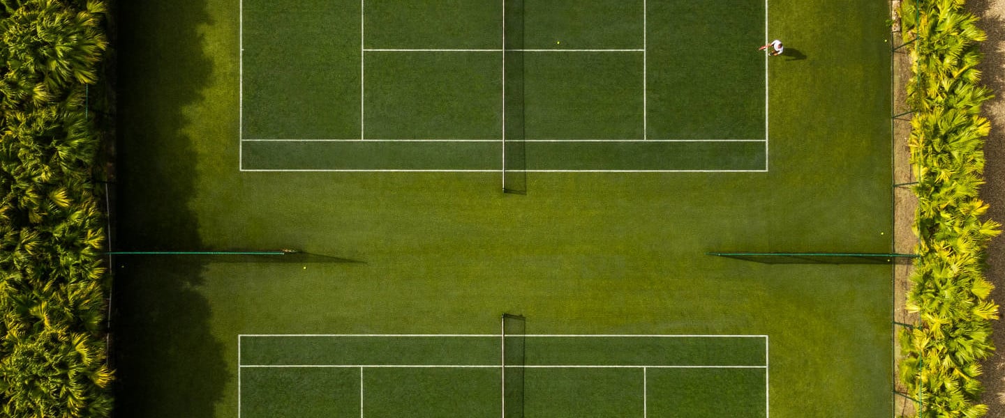 Overhead view of four tennis courts with white lines on manicured grass, bordered by vibrant hedges and natural landscape.