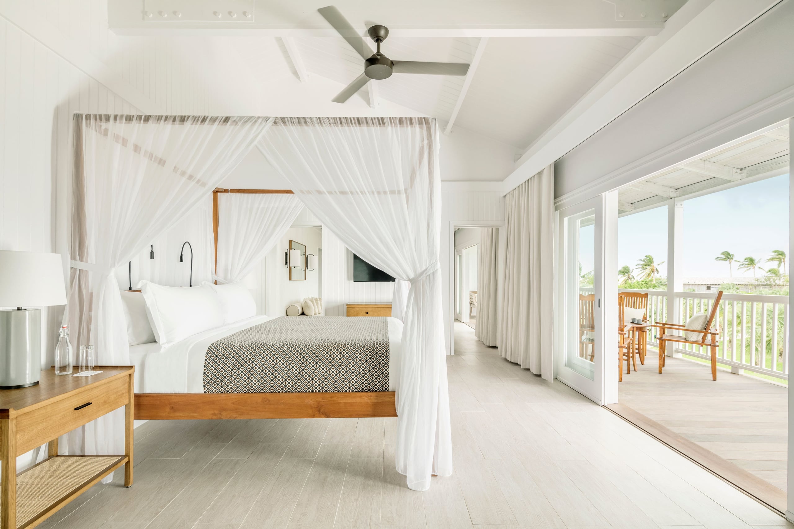 COMO Parrot Cay, North America — Coastal bedroom with wooden four-poster bed, sheer white canopy, ocean-view terrace, ceiling fan, and minimalist modern furnishings.