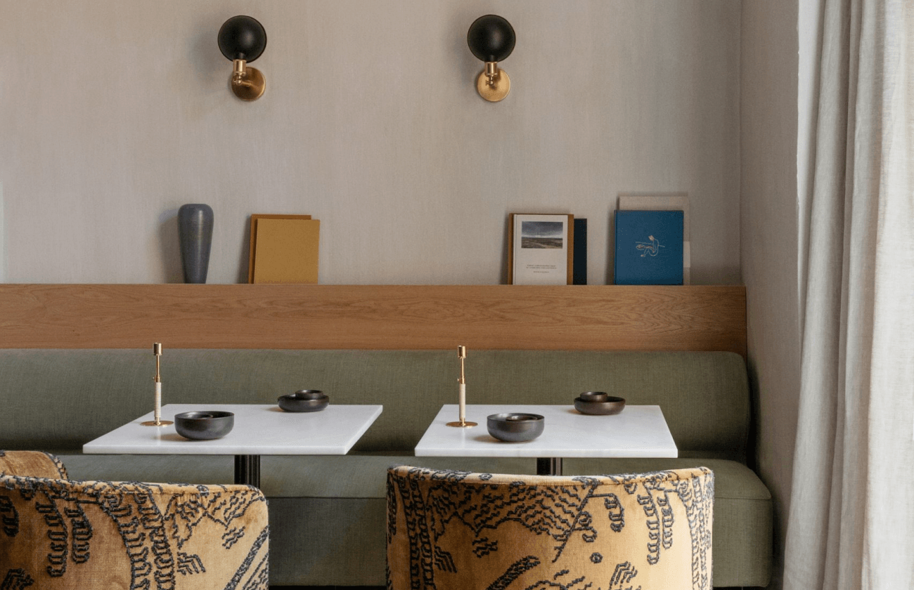 The Audo Salon — Audo Salon, Europe — Banquette seating with sage upholstery, white tables, brass sconces, and curated books against neutral walls, modern hospitality interior design.