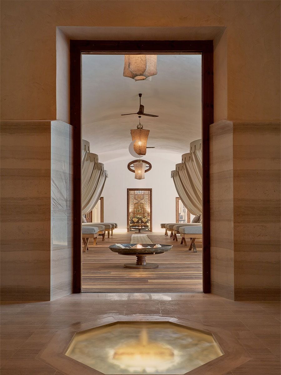 Cap Rocat, Balearic Islands — Ochre corridor opening onto minimalist lounge with woven pendant lights, ceiling fan, linen curtains, and central fire bowl.