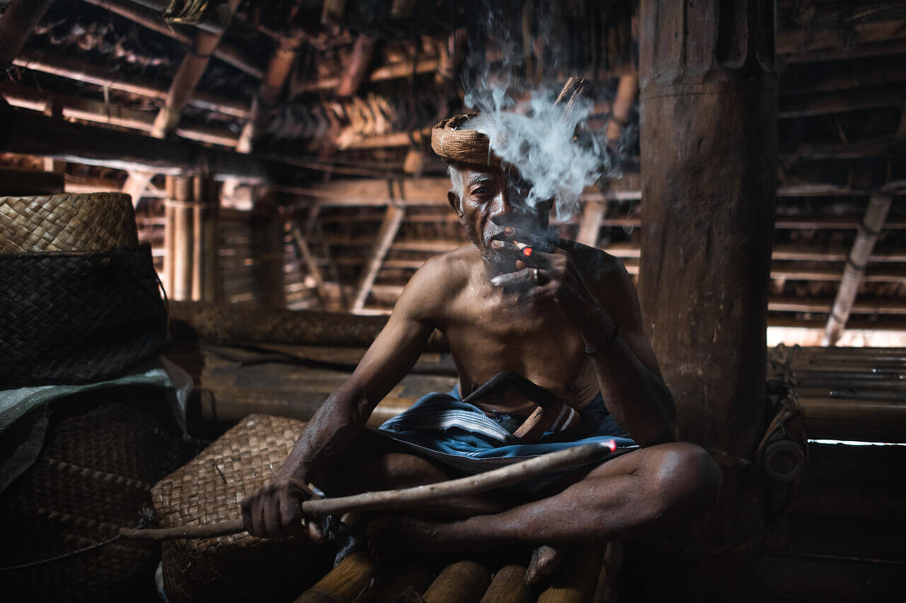 Cap Karoso, Asia — Shirtless man smoking cigar in wooden interior, dappled sunlight through thatch roof, rustic vernacular architecture, atmospheric tropical lodge setting.