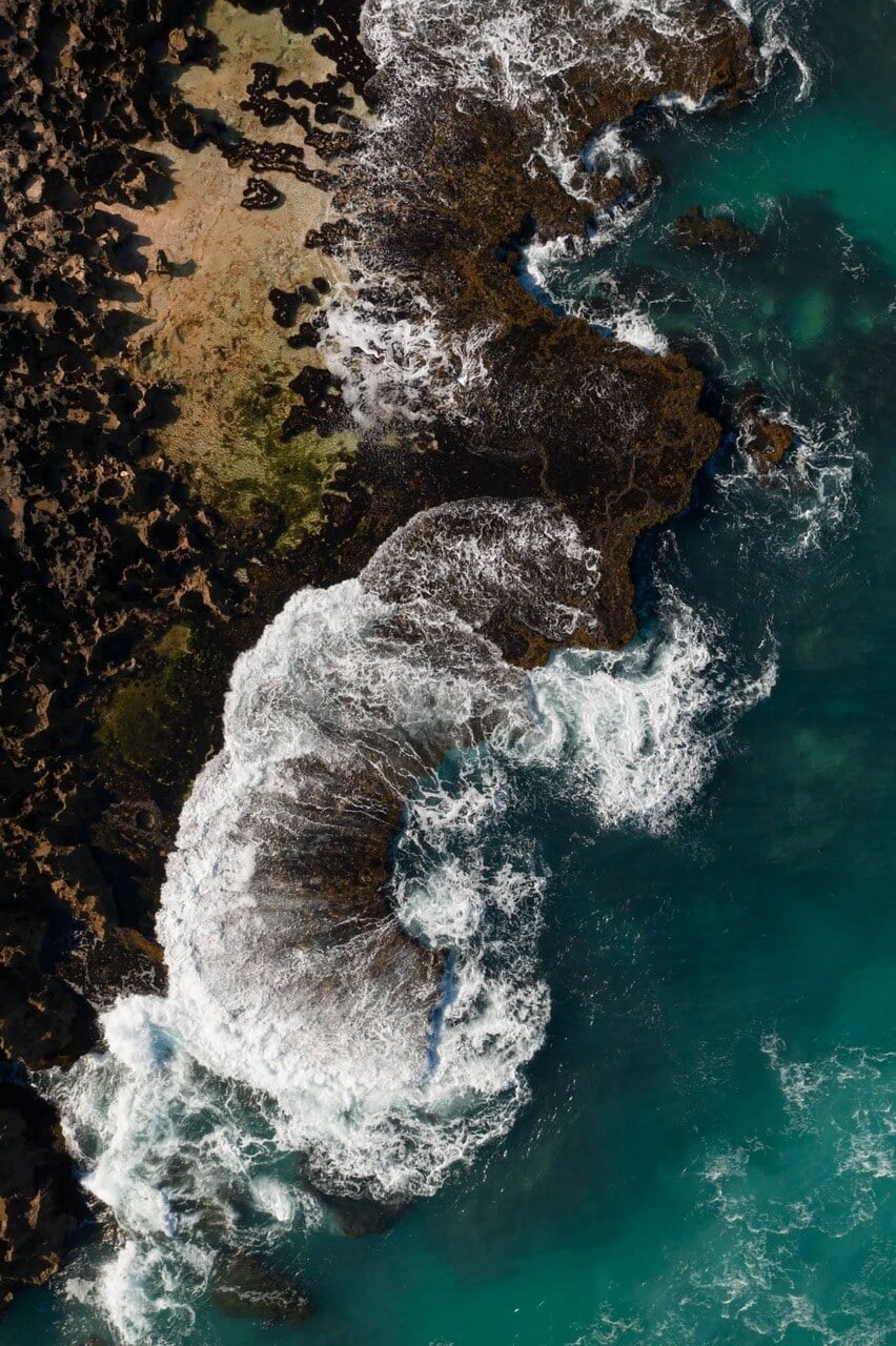 The Wild — Aerial view of rocky coastal cliffs with turquoise ocean and white wave patterns, luxury travel destination aesthetic.