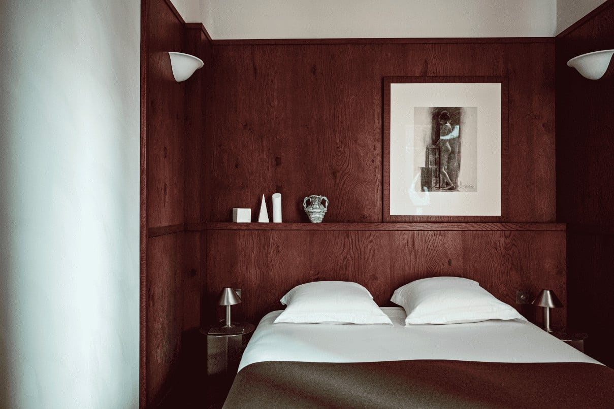Académies et des Arts, Europe — Deep burgundy wood-paneled bedroom with floating shelf, white bedding, minimalist decor, and framed artwork above bed.