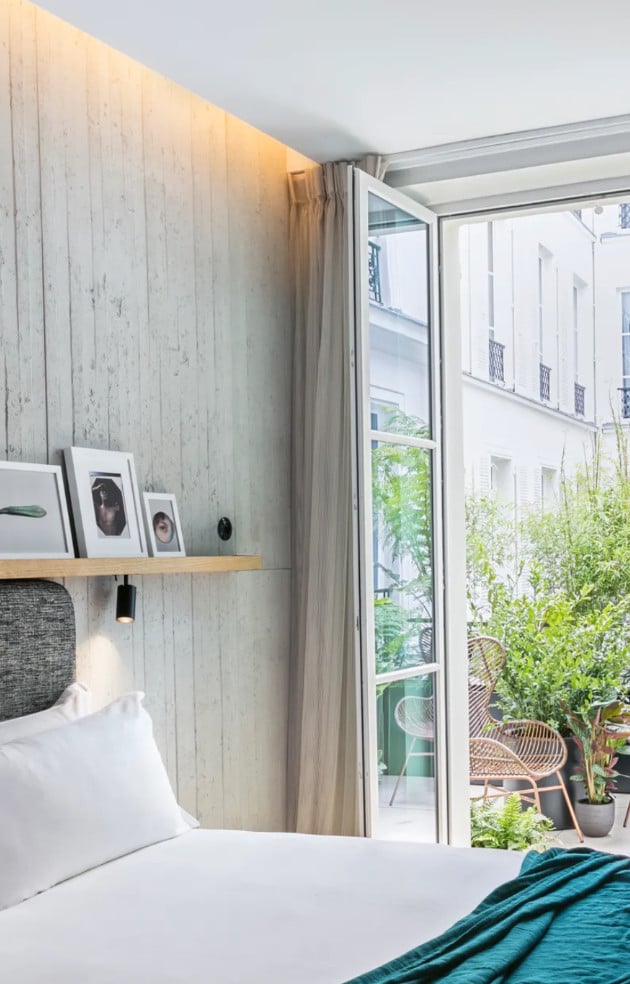 Hôtel National Des Arts, Europe — Modern bedroom with whitewashed wood wall, floating shelf, pendant lighting, floor-to-ceiling glass doors opening to verdant balcony terrace, Parisian architecture beyond.