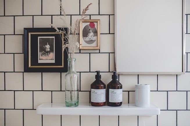 Le Barn, Europe — Minimalist white shelf against subway tile wall displaying vintage portraits, clear glass vase with dried botanicals, amber apothecary bottles, and white candle in serene luxury interior.