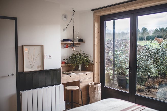 Le Barn, Europe — Minimalist bedroom with black-framed glass doors opening to countryside, natural wood furniture, white radiator, potted plants, and soft natural light.