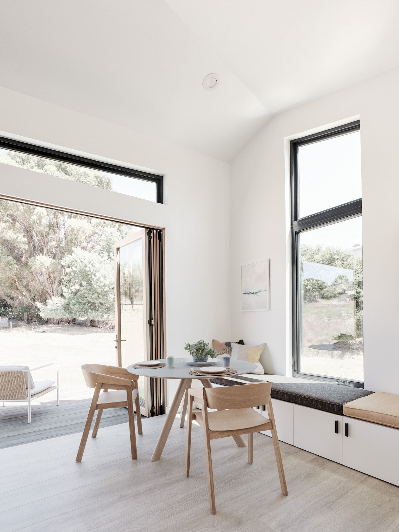 Abodu x Koto, North America — Minimalist dining nook with natural light, wooden chairs, round table, and built-in window seat overlooking landscaped grounds.