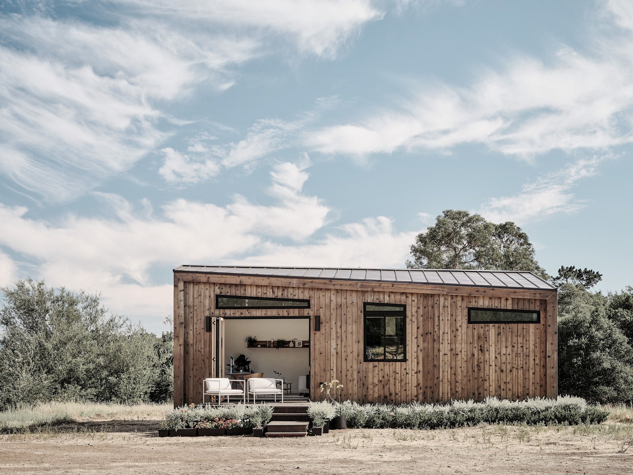 Abodu x Koto, North America — Modern timber cabin with board-and-batten siding, steel roof, floor-to-ceiling windows, minimalist interior, surrounded by native desert landscaping under blue sky.
