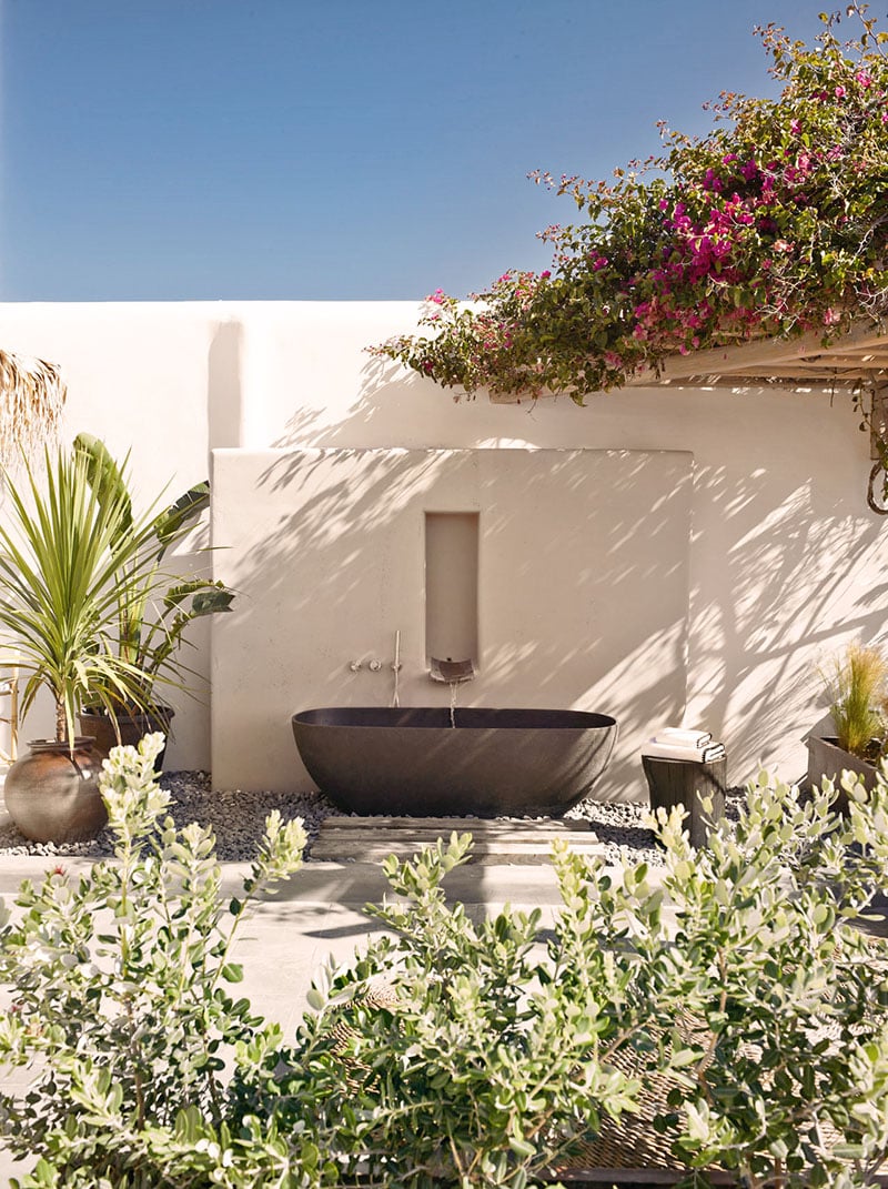 The Wild, Europe — Minimalist outdoor bathroom with dark stone soaking tub against white walls, magenta bougainvillea overhead, Mediterranean garden plants and clear sky.