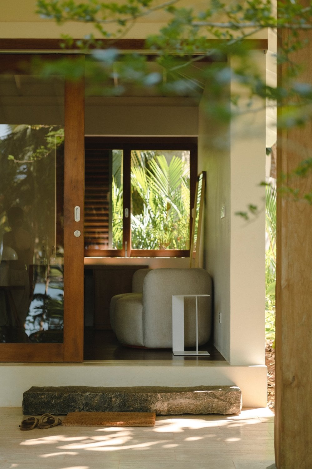 Kurulu Bay, Asia — Minimalist villa entrance with wooden sliding doors, curved stone seating, palm views, natural light filtering through tropical foliage.