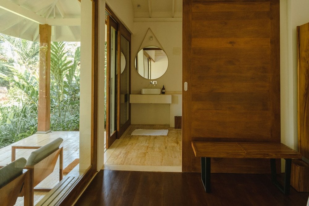 Kurulu Bay, Asia — Minimalist bathroom gallery with teak wood accents, round mirror, natural light from garden views, and wooden bench seating.
