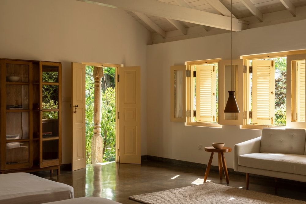 Kurulu Bay, Asia — Minimalist bedroom with natural light through louvered windows, concrete flooring, linen sofa, wooden furnishings, and open doors revealing tropical garden views.