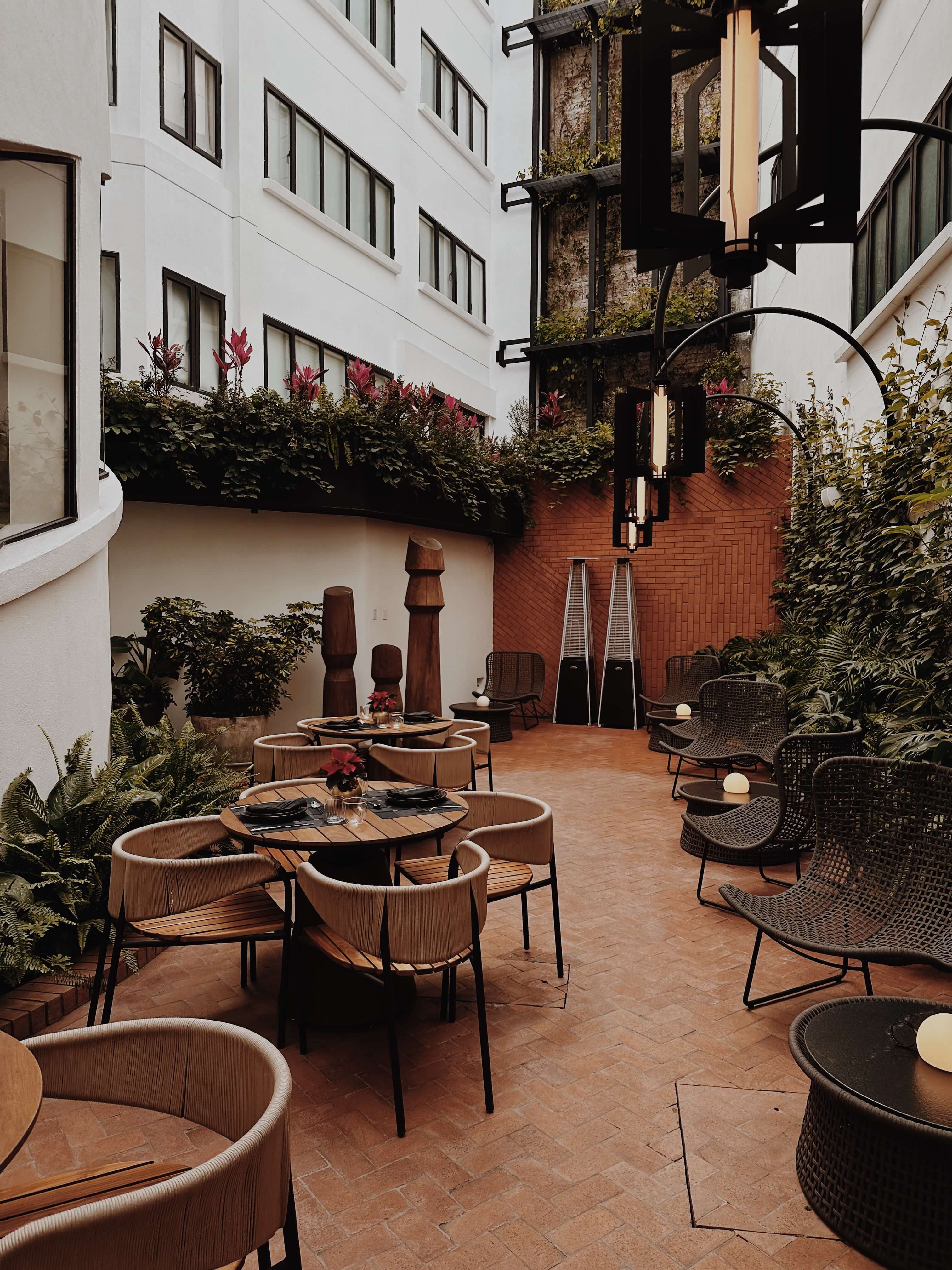 Campos Polanco, Mexico City, Mexico — Courtyard dining space with terracotta tile flooring, woven lounge chairs, wooden tables, brick walls, climbing ivy, and modern sculptural planters.