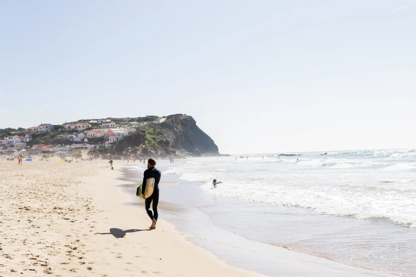 Costa Vicentina Surfing — Surfer in black wetsuit carrying yellow surfboard walking into Atlantic waves at coastal cliff village, golden hour light.