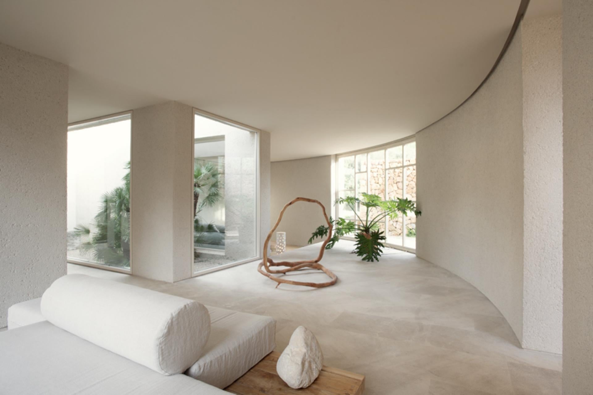 Es Racó d'Artà, Balearic Islands, Spain — Minimalist curved interior with polished concrete floors, floor-to-ceiling windows, sculptural wooden chair, and potted palms overlooking courtyard.