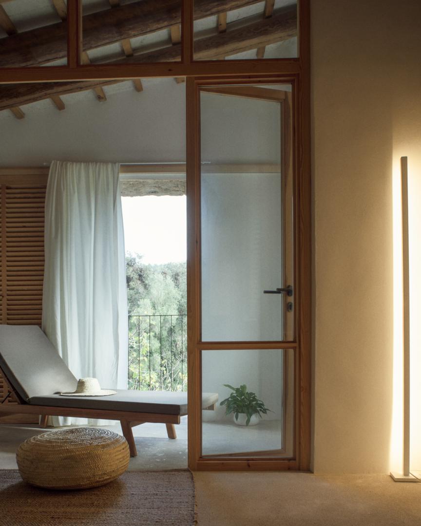 Es Racó d'Artà, Balearic Islands, Spain — Minimalist bedroom with wooden-framed glass doors opening to forested hillside views, cream linen curtains, mid-century furniture, warm terracotta walls.