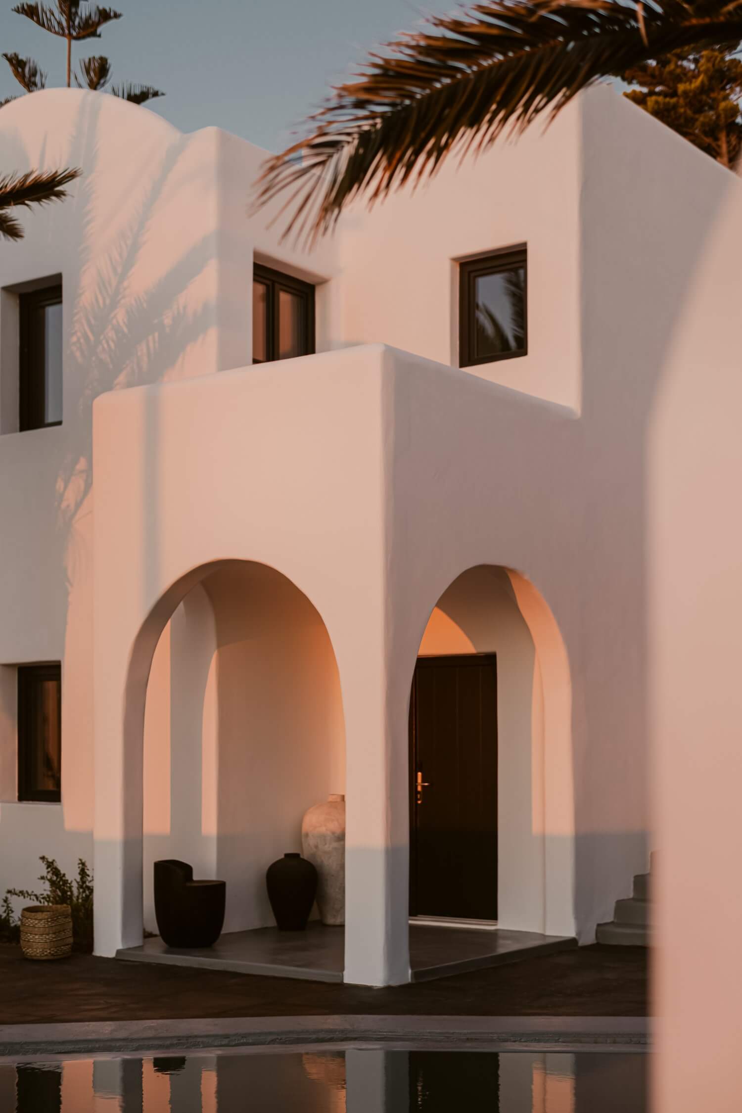 Nobu Santorini, Greece — White-washed architecture with curved arches, warm golden-hour light, palm fronds, dark wooden door, minimalist luxury Mediterranean design.