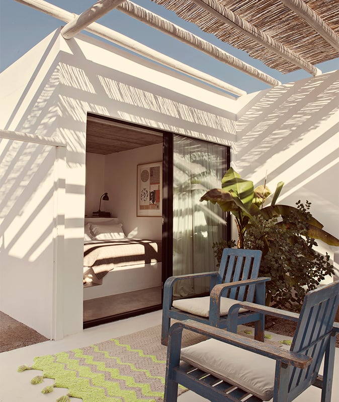 Pensão Agrícola, Europe — Minimalist bedroom terrace with woven pergola, blue lounge chairs, potted plants, and dappled shadow patterns on white walls.