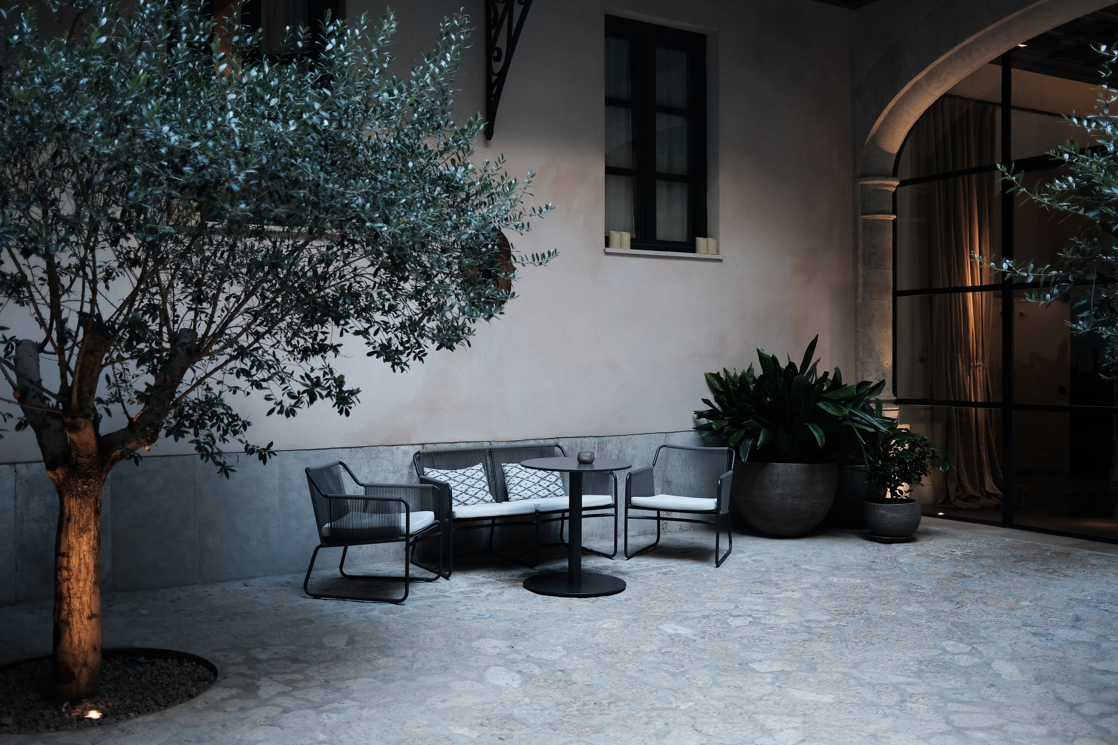 Sant Francesc Hotel Singular, Balearic Islands, Spain — Minimalist courtyard with black lounge seating, olive tree, potted plants, arched colonnade, and concrete flooring at dusk.