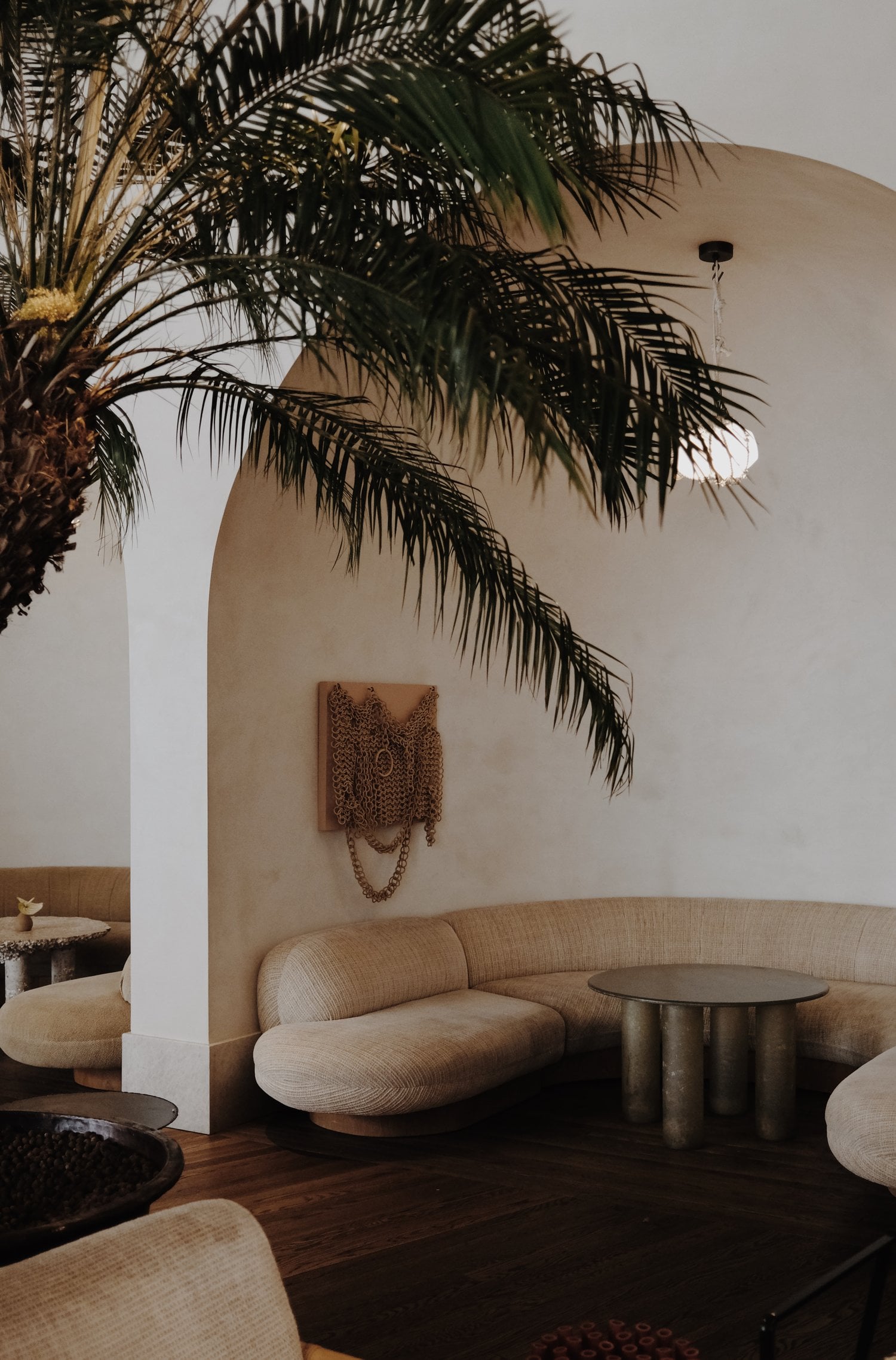 Santa Monica Proper, California, United States — Curved cream sofa beside metal table beneath arched wall, palm fronds casting shadows, minimalist luxury lounge.